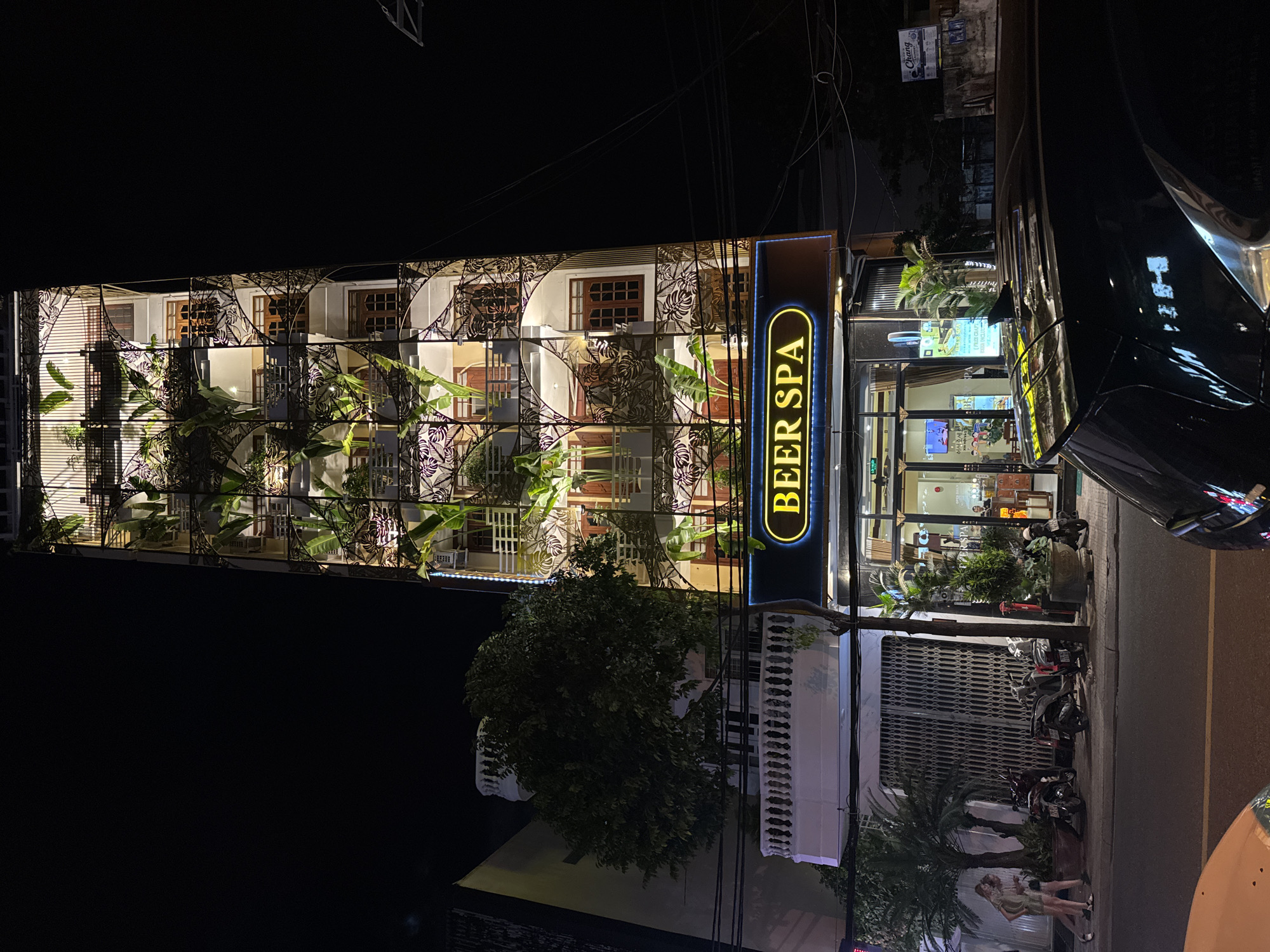 The image shows a tall, multi-story building at night, identified by a lit sign as "BEER SPA". The building's facade is decorated with vertical planters containing green foliage, creating a tiered garden effect. The building has multiple windows and balconies with decorative metalwork. A large tree is visible to the left of the building. In the foreground, a street is visible with parked motorcycles and two individuals walking on the sidewalk. A black car is parked in the lower right corner. The scene appears to be in a city, with streetlights and building lights illuminating the area. The architecture and presence of palm trees suggest a tropical or sub-tropical location. The sign "BEER SPA" in English, along with the general aesthetic, is consistent with commercial establishments in tourist areas of Nha Trang, Vietnam.