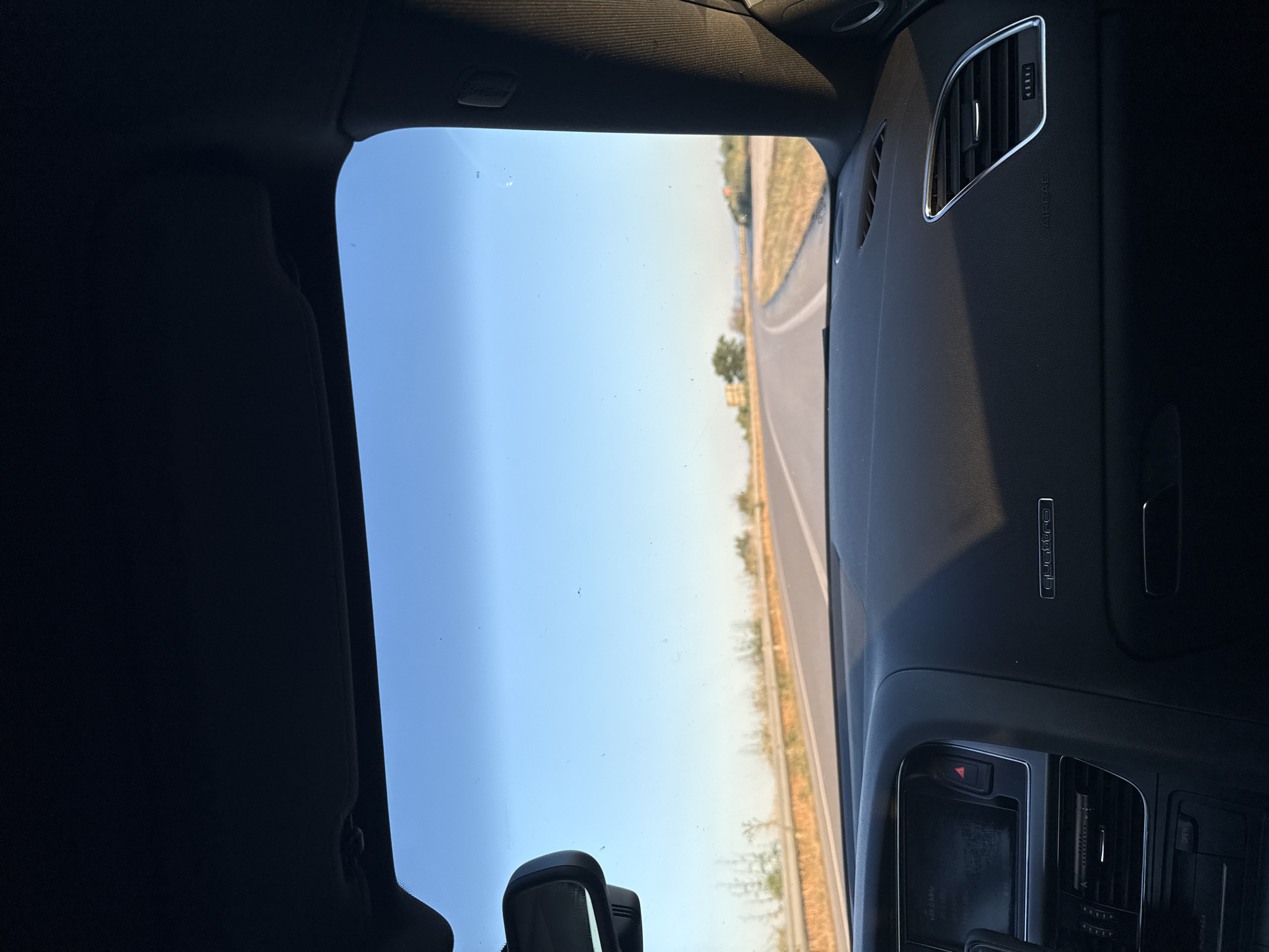 The image captures a view from inside a car, looking out through the windshield. The car's dashboard is visible in the foreground, with a central console featuring a screen and various controls. To the right of the console, the word "quattro" is embossed on the dashboard. The windshield displays a clear, light blue sky with a visible road stretching into the distance. A guardrail runs along the side of the road, separating it from dry, grassy terrain with sparse vegetation and a few trees. The perspective suggests the car is in motion on a highway. The scene appears to be set in a rural or suburban area under bright daylight. The location is noted as Unknown, Greece.