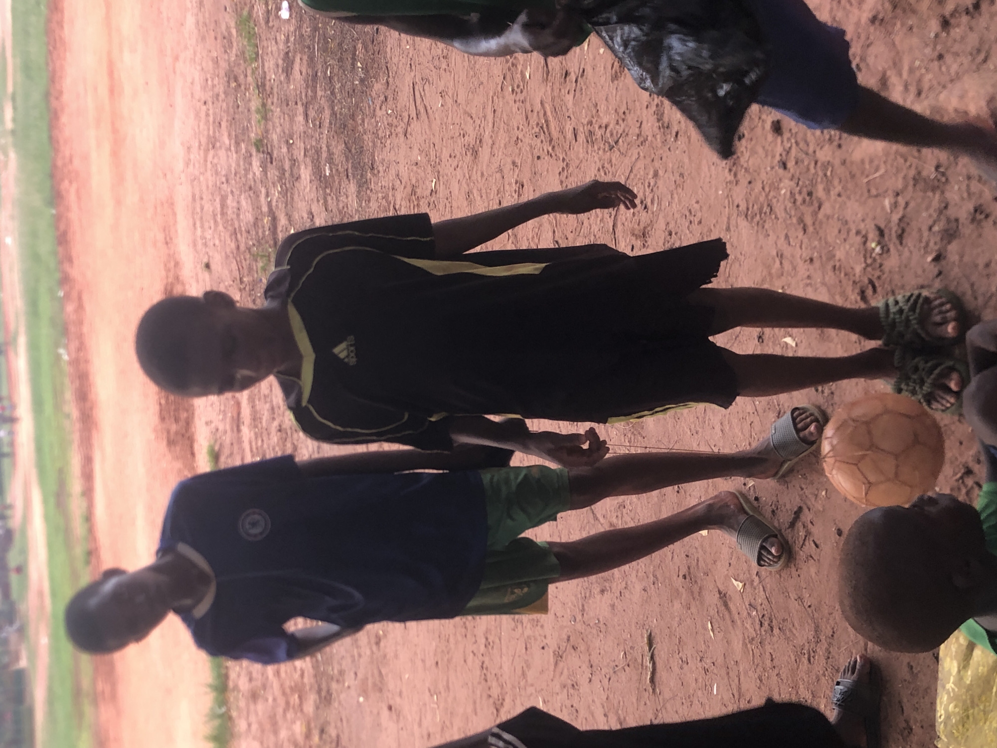 The image shows a group of people standing on a dirt field in Nsugbe, Nigeria. Two boys wearing dark t-shirts and shorts are standing near a soccer ball on the right side of the frame. Other people can be seen in the background. The field appears to be adjacent to a grassy area, possibly a sports field. The dirt is a reddish-brown color. The individuals appear to be interacting near a soccer ball. They are wearing sandals. One boy is wearing an Adidas shirt.