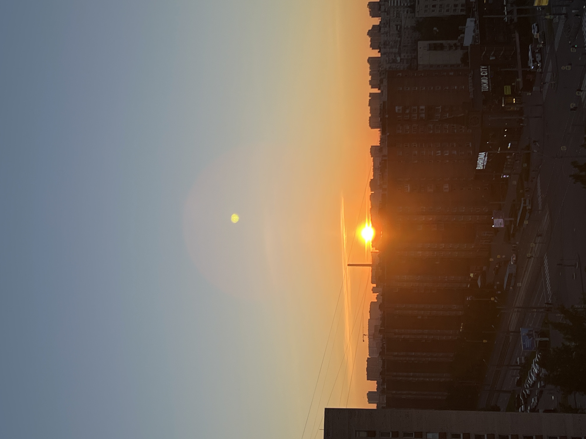 The image captures a sunset over a dense urban landscape in Sinyavino, Russia. The sky transitions from a pale blue at the top to a vibrant orange and yellow near the horizon, indicating the sun is low. A bright, circular sun is visible just above the skyline, casting a warm glow and creating lens flare effects in the sky. In the mid-ground, a series of multi-story apartment buildings form a continuous, dark silhouette against the illuminated sky. Signs for "BAHROMA" and "TOKIO-CITY" are partially visible on one of the buildings. Below the buildings, a street is lined with parked cars and a bus. Power lines crisscross the scene, and a distant construction crane is visible. The overall impression is of a typical evening in a residential area of a Russian city.