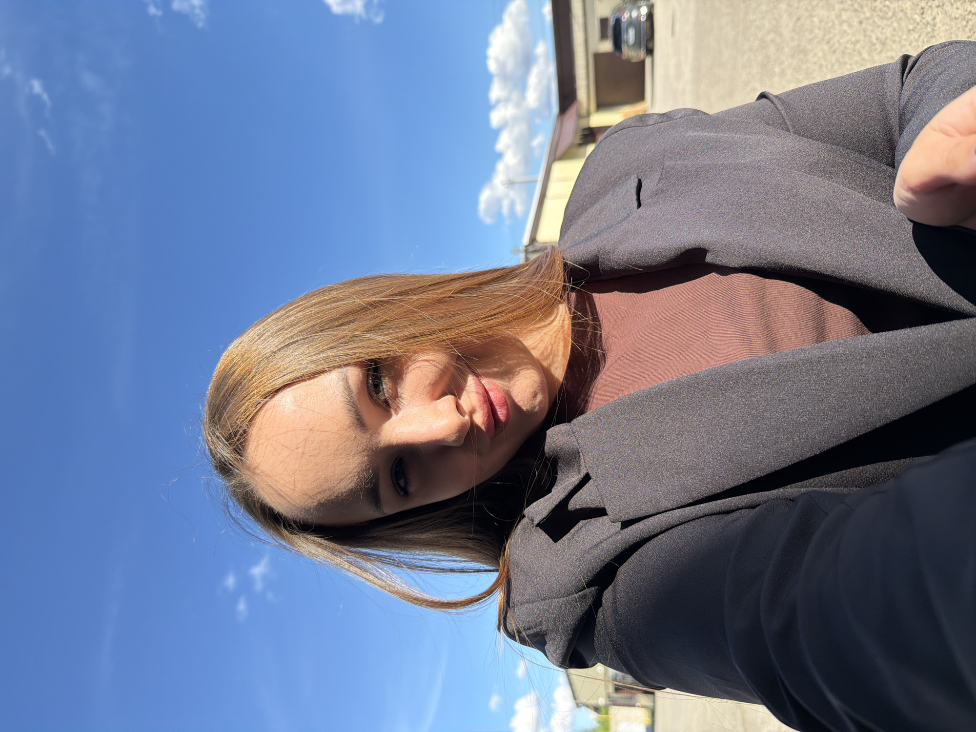 A woman with light brown hair is facing the camera in an outdoor setting under a clear blue sky with scattered white clouds. She is wearing a brown shirt and a black blazer. The background includes a building with a car parked nearby. The location is likely Chaikovsky, Russia, as indicated by the prompt. The woman appears to be taking a selfie.