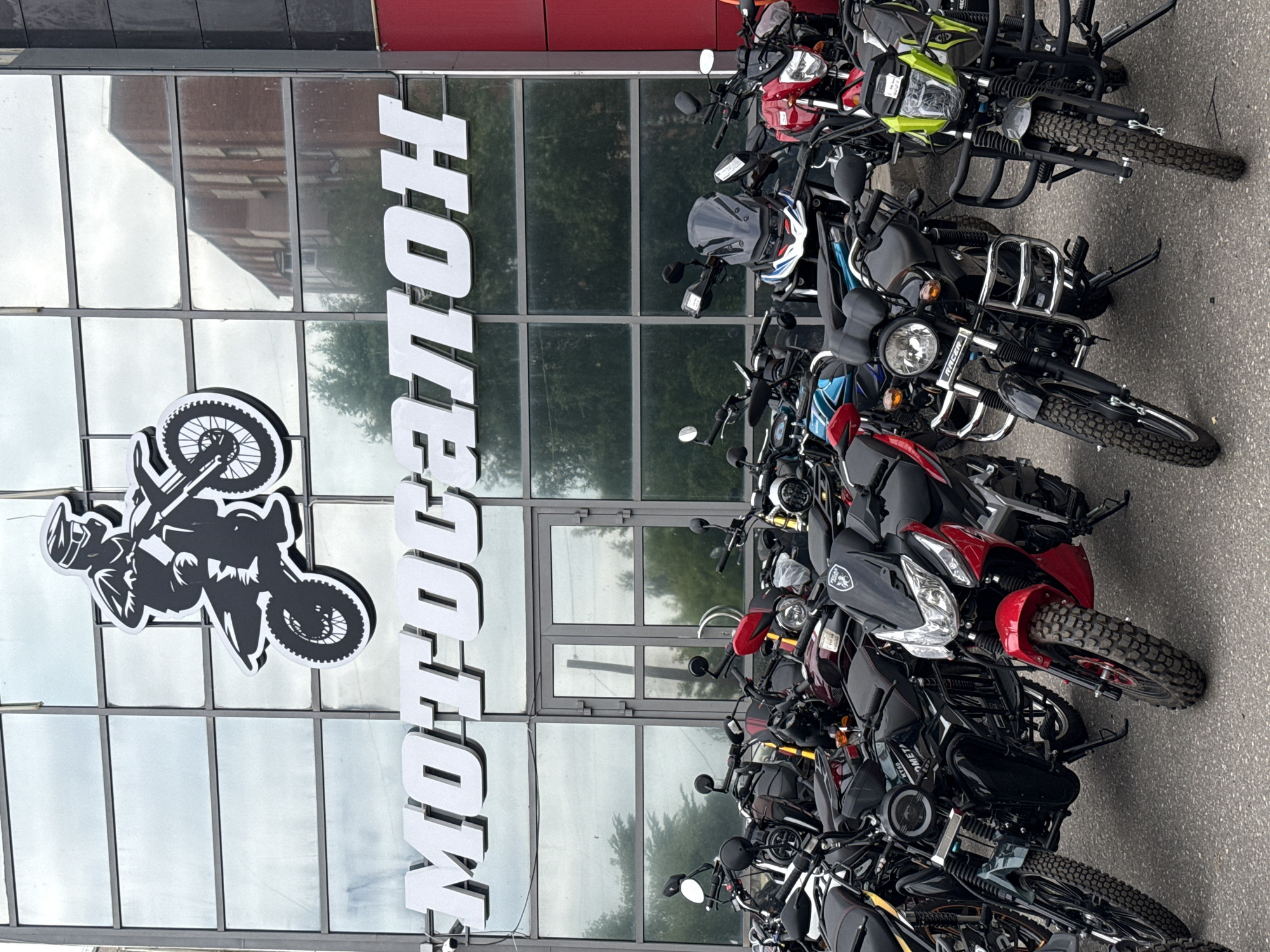 The image shows a motorcycle dealership called "MOTOCABOH" in Votkinsk, Russia. The dealership has a large glass facade with the name "MOTOCABOH" prominently displayed in white lettering. Above the text, there is a black silhouette of a motorcyclist riding a dirt bike. In front of the dealership, a variety of motorcycles are parked in rows. The motorcycles are of different colors and styles, including street bikes and off-road bikes. The environment appears to be an urban setting with trees and buildings visible in the background, reflected in the dealership's windows. The overall scene suggests a commercial establishment focused on the sale of motorcycles.