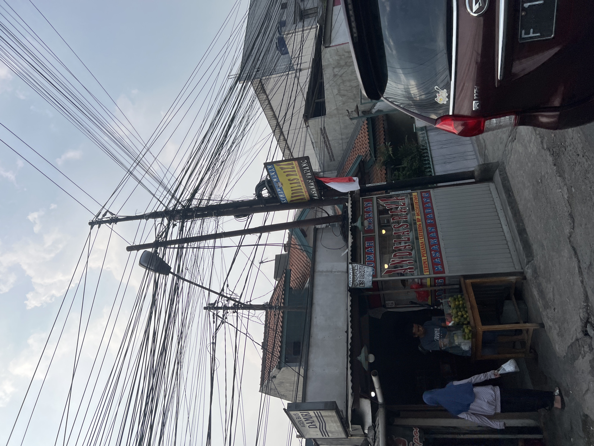 In the city of Citeureup, Indonesia, an outdoor scene unfolds with a prominent display of chaotic electrical wires crisscrossing the sky. Dominating the foreground is a maroon car, its rear visible, bearing a license plate starting with "F 1". Adjacent to the car is a food stall with a sign that reads "RUMAH MAKAN ANDALAS RAJA" and "MASAKAN PADANG". An Indonesian flag hangs from a pole near the stall. Two individuals are present at the stall; one person, wearing a blue headscarf and a light-colored patterned jacket, walks away carrying a white plastic bag, while another person, dressed in a grey t-shirt, stands behind a table laden with oranges. The stall is situated next to a building with a sign that says "AGEN BUS DAMRI". The sky is overcast with visible clouds.