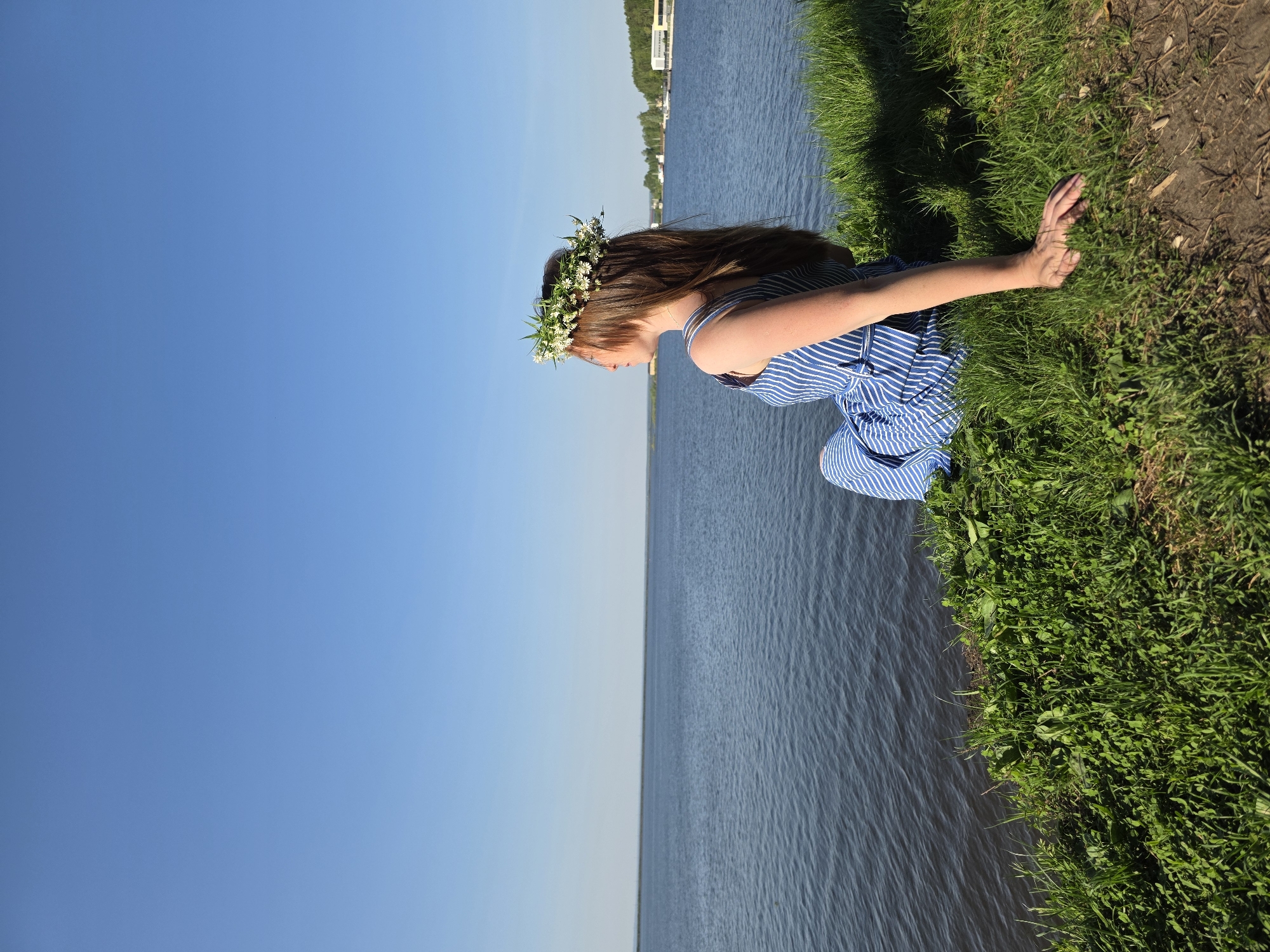 A woman with long brown hair, wearing a blue and white striped dress and a flower crown, is kneeling by the edge of a body of water. She has her right arm extended and her hand is resting on the green grass. The water body appears to be a river, possibly the Kama River. The location could be Chaikovsky, Russia, based on the prompt. The sky is clear blue. In the distance, on the opposite side of the water, there are some buildings. The area around the kneeling woman has green grass and some bare earth.