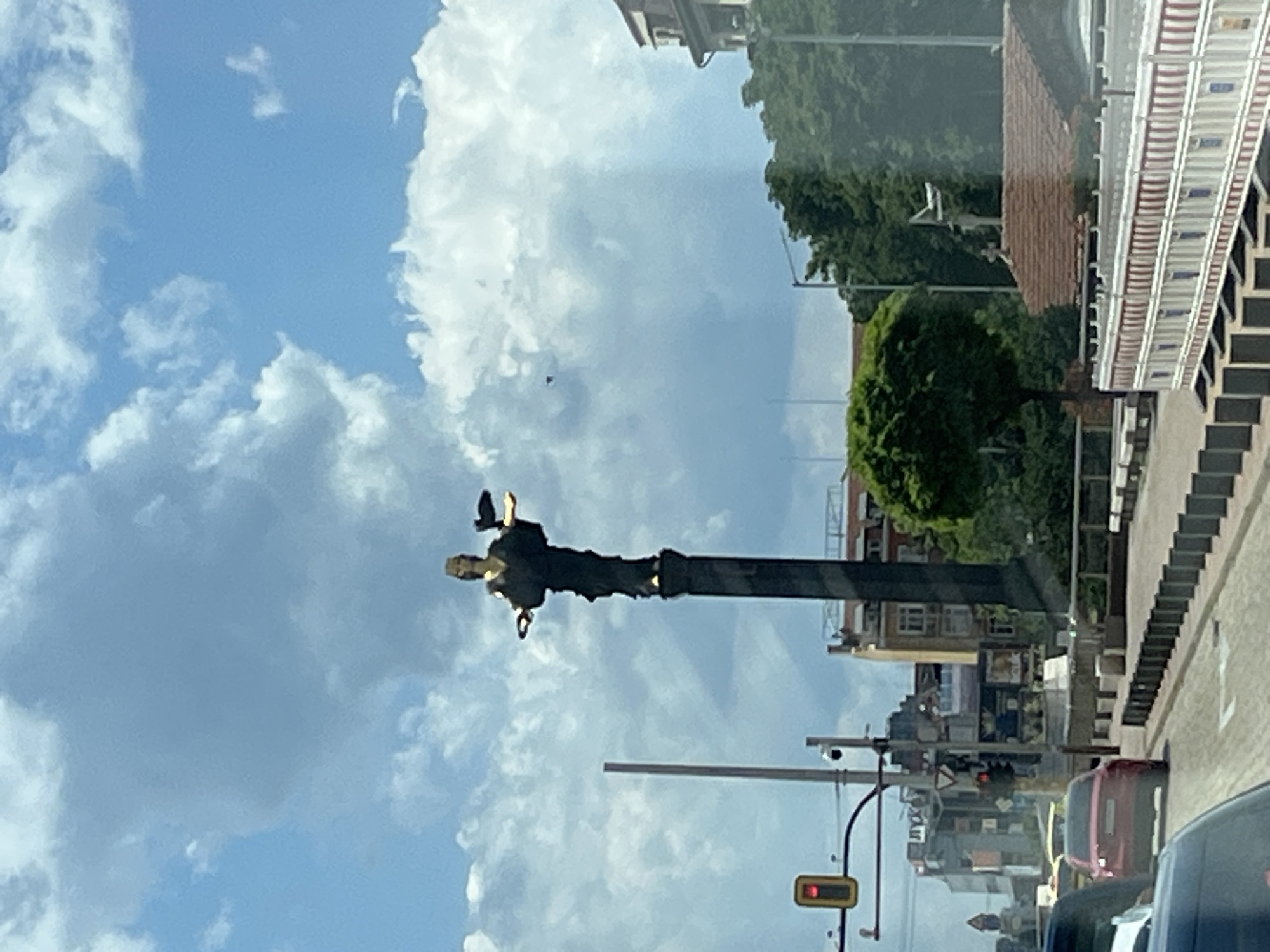 This media file depicts a street scene observed from a vehicle. The primary subject is a tall, dark monument or statue standing on a pillar, with what appears to be a figure at its apex. The monument is set against a backdrop of a bright blue sky with fluffy white clouds. To the right of the monument, there are buildings with red brick and windows, and a sidewalk with stairs. Trees are visible behind the buildings, suggesting an urban or suburban setting. Further to the right, a road is visible with cars and a traffic light. The overall impression is that of a sunny day, possibly late afternoon given the lighting. The image is taken from inside a vehicle, as indicated by the reflection on the surface and the perspective. The image is oriented vertically.