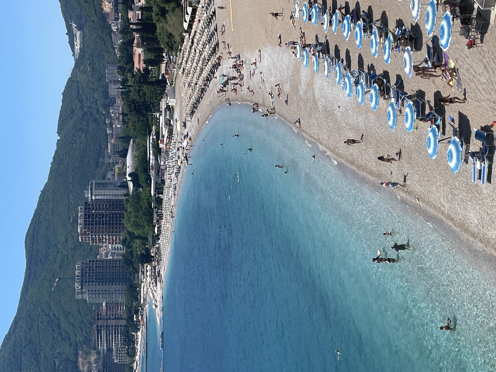This aerial shot captures the Mogren beach area within the coastal city of Budva, Montenegro. The frame showcases a curved shoreline, where the clear turquoise waters of the Adriatic Sea meet the sandy beach. A densely populated area can be seen enjoying the beach, with numerous beachgoers sunbathing, walking along the shore, and swimming in the sea. Parallel rows of blue and white sun umbrellas are arranged closely together, casting shadows on the sand. Dense green vegetation covers the mountain rising to the left of the image, with buildings and other structures interspersed among the trees. Several tall residential and commercial buildings are along the coast, adjacent to the beach, and are visible from the water's edge.