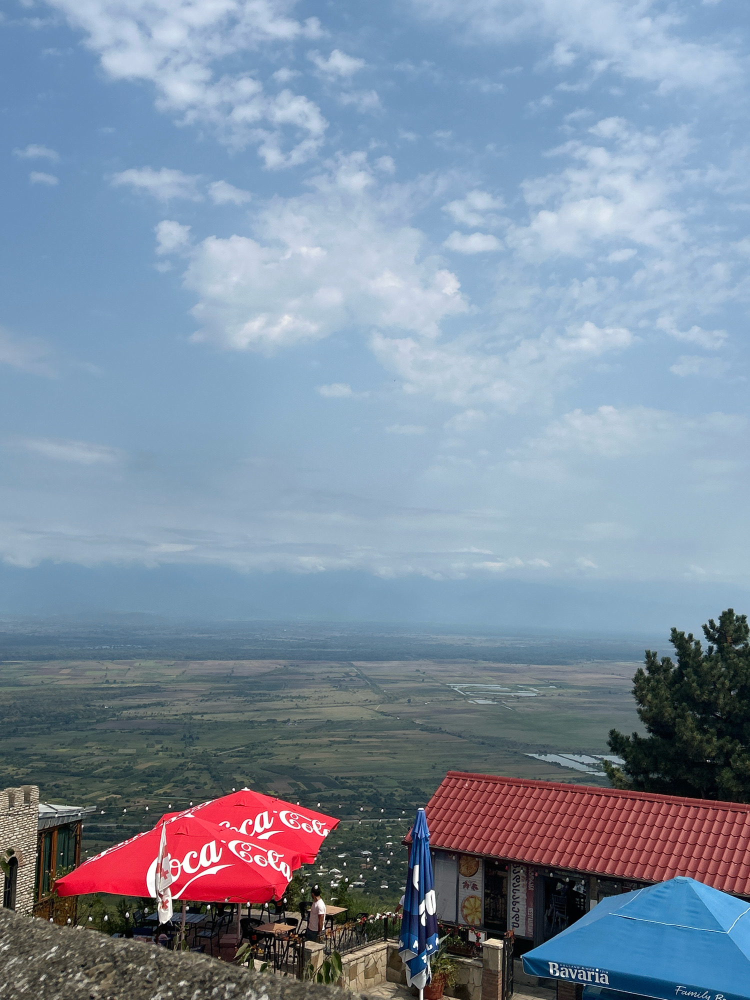 The image depicts an outdoor seating area of a restaurant or cafe, offering a panoramic view of a valley and distant mountains. In the foreground, several umbrellas provide shade. Two large red umbrellas with the Coca-Cola logo are prominent, while a blue umbrella with the Bavaria logo is visible on the right. A red-tiled roof of a building is also visible. The background showcases a sprawling landscape of fields, with a river or body of water winding through it. The sky is partly cloudy, with a mix of blue sky and white clouds.
The location is identified as Sighnaghi, Georgia. Sighnaghi, often referred to as the "City of Love," is a charming town located in the Kakheti region of Georgia. It is known for its well-preserved 18th-century architecture, cobblestone streets, and stunning views of the Alazani Valley and the Greater Caucasus Mountains. The town is a popular tourist destination, attracting visitors with its historical significance, wine culture, and picturesque setting. The view from this vantage point suggests it is likely from a restaurant or cafe situated on a hillside overlooking the valley.