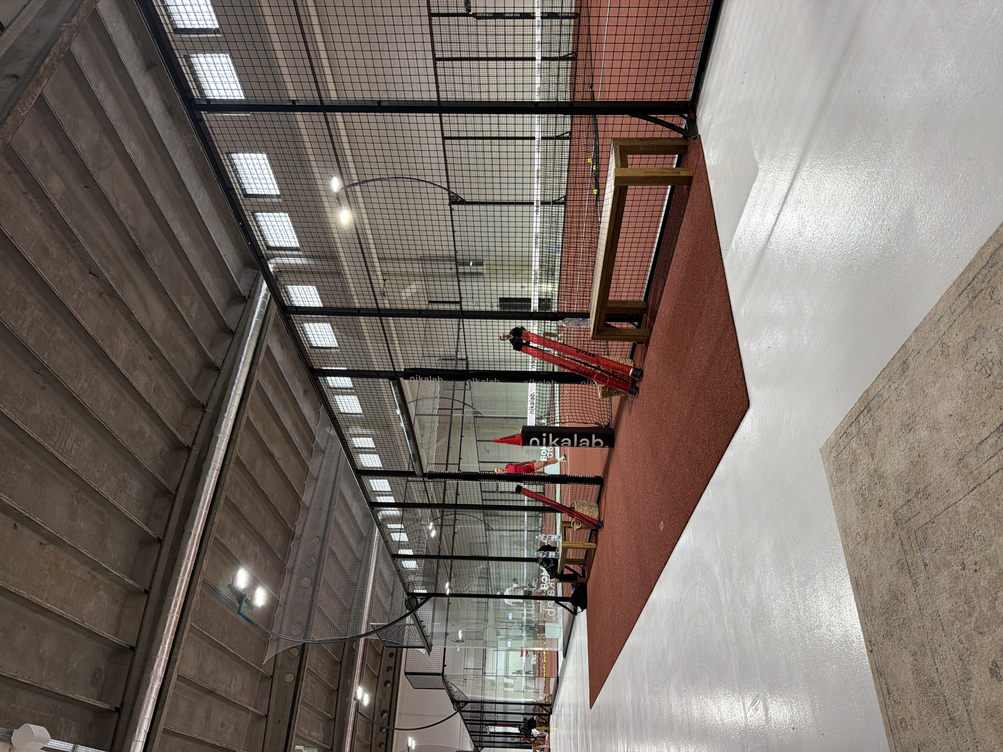 The image depicts the interior of a padel tennis facility, presumably in Unknown, Unknown. The court is enclosed by wire mesh fencing. The floor is divided into a reddish-brown playing surface and white adjacent areas. Near the front of the court are benches and equipment, including red objects and black bags. Advertising for "nikalab" is visible. The ceiling has a grid of light fixtures and is supported by concrete beams. Some people are on the tennis court playing padel.