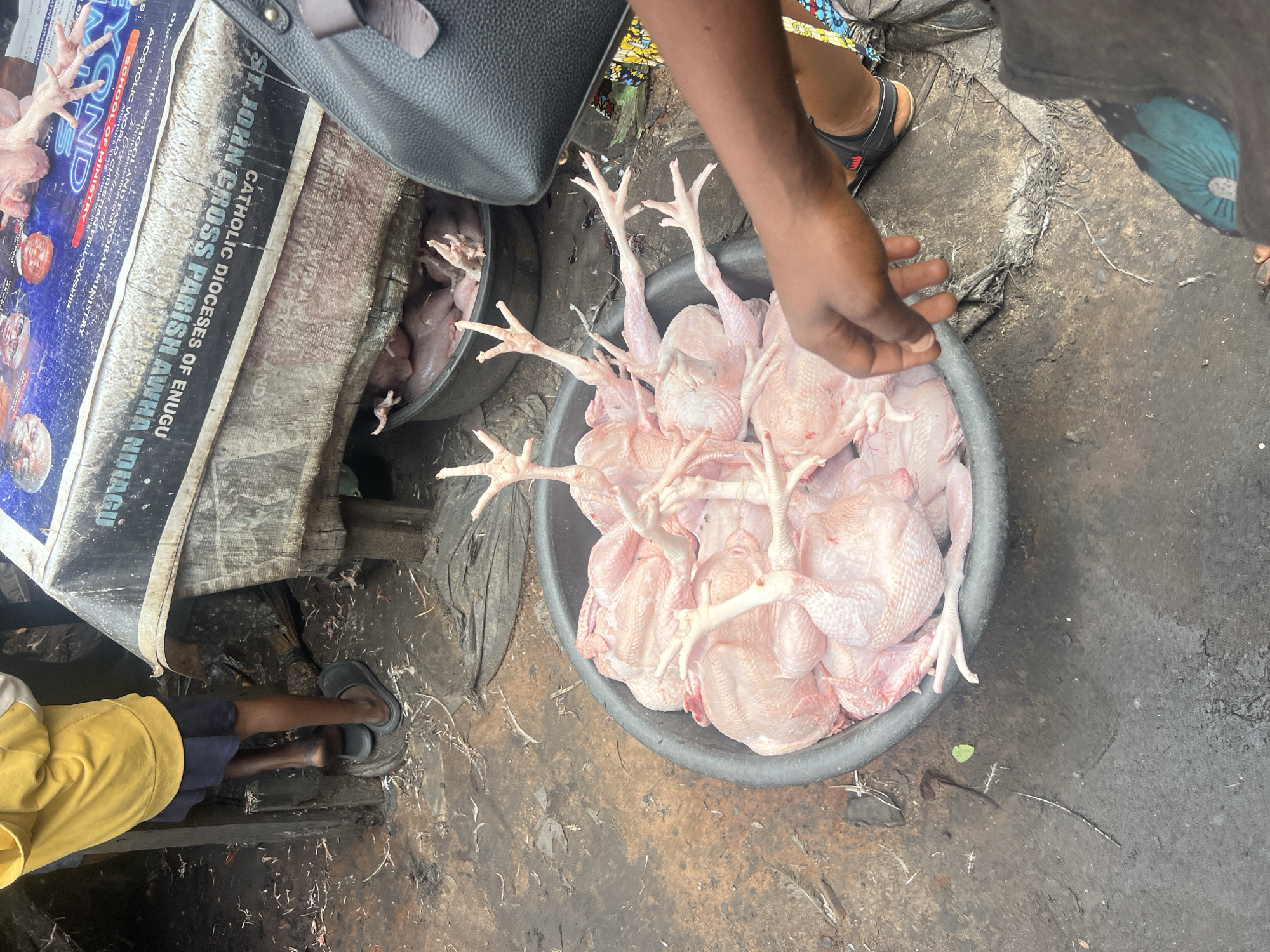 In Nkpor, Nigeria, a close-up shot captures a street market scene where an individual, identified by their dark skin tone and casual attire, is interacting with a large black basin filled with raw chickens. The chickens, pale pink and unplucked, are arranged with their legs and feet prominently displayed. The environment appears to be an outdoor market, with a dirty, wet concrete ground surface and a rustic wooden structure in the background. A dark-colored handbag rests on the edge of the scene, and a piece of cloth with a vibrant blue and yellow pattern is visible. The overall impression is one of a bustling local marketplace, focusing on the sale of fresh produce.