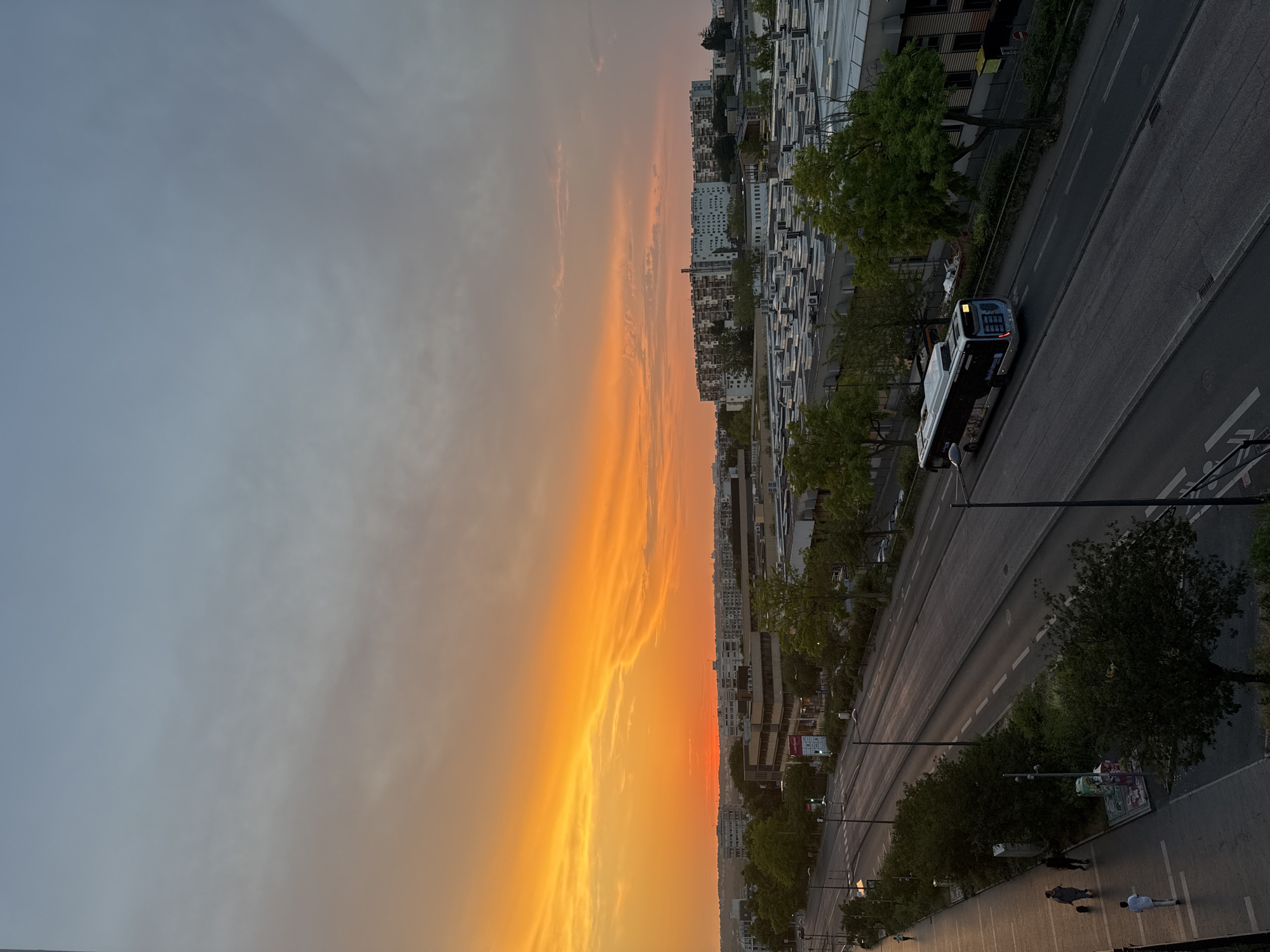 A wide shot captures a city street at dusk, with a vibrant orange and yellow sunset dominating the sky. The horizon is lined with buildings, some appearing to be apartment complexes and others industrial structures with flat, white roofs. A multi-lane road runs horizontally through the mid-ground, with a single white and black bus driving on the right side. On the left side of the road is a pedestrian walkway with a few individuals walking. Trees line both sides of the road and walkway, their leaves a deep green. The sky transitions from the intense sunset colors at the horizon to a pale blue and gray at the top of the frame. The overall scene is one of urban life at the close of day.