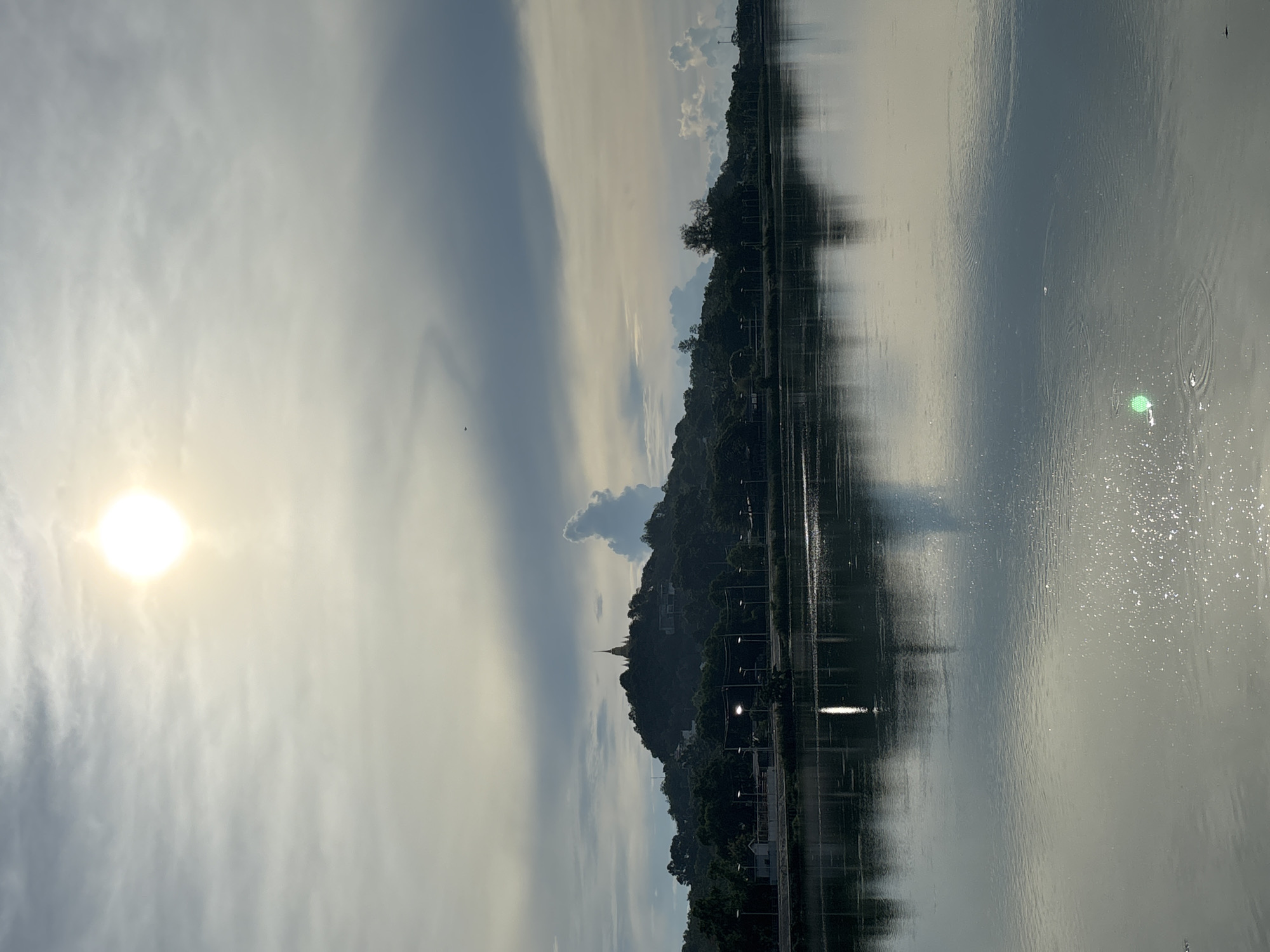 The image shows a daytime scene in บ้านบางรักษ์, Thailand. A body of water, possibly a lake or river, dominates the right side of the image. Reflections of the opposite bank are visible in the water. On the left, a landscape is present, including a hill with trees and a building structure possibly a temple or stupa at its peak. A road with streetlights runs along the base of the hill. The sky features a bright sun on the left and a mix of clouds.