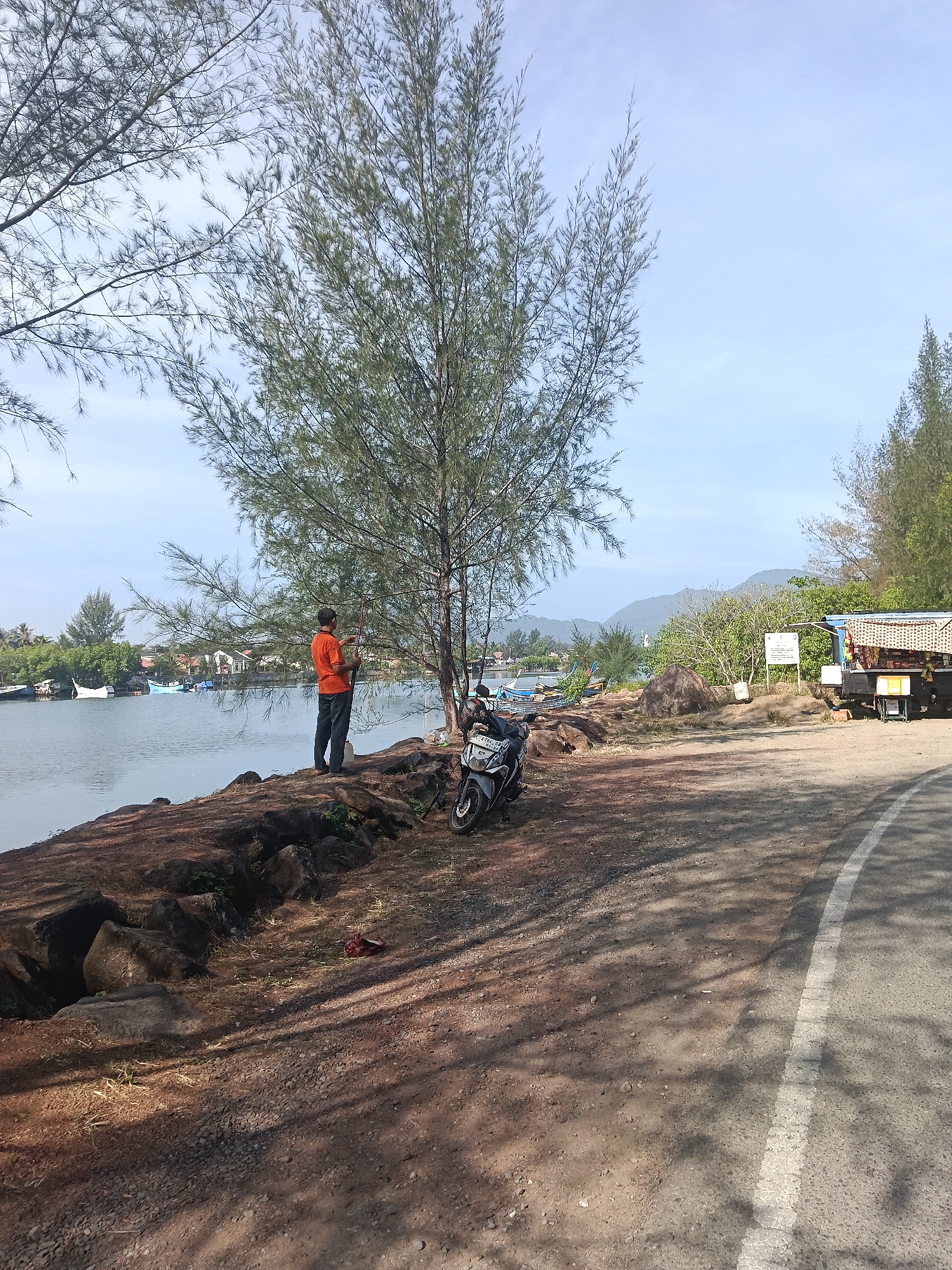 The image depicts a man in an orange shirt and dark pants standing by a body of water in Banda Aceh, Indonesia. He is holding a fishing rod, suggesting he is fishing. A motorcycle is parked on the rocky shore next to him. In the background, there are boats on the water, buildings, and lush green hills under a clear blue sky. The scene appears to be a coastal or riverine area within the city.