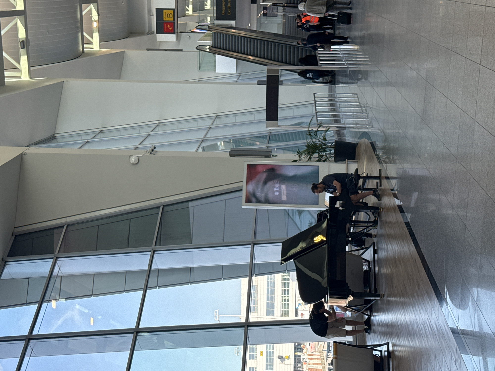 The image depicts the interior of Brussels Airport in Zaventem, Belgium. A grand piano is set up in the main concourse, with a pianist seated and playing. To the left of the piano, another individual is bent over, seemingly adjusting or preparing the instrument. Sunlight streams in through large, angled windows that offer a view of the airport tarmac, with buildings and aircraft visible in the distance. In the background, escalators and signage indicating "Gates B" and "Baggage / Exit Brus" are visible, along with other travelers and airport staff moving through the space. The floor is a combination of polished wood and tile, reflecting the bright interior lighting.