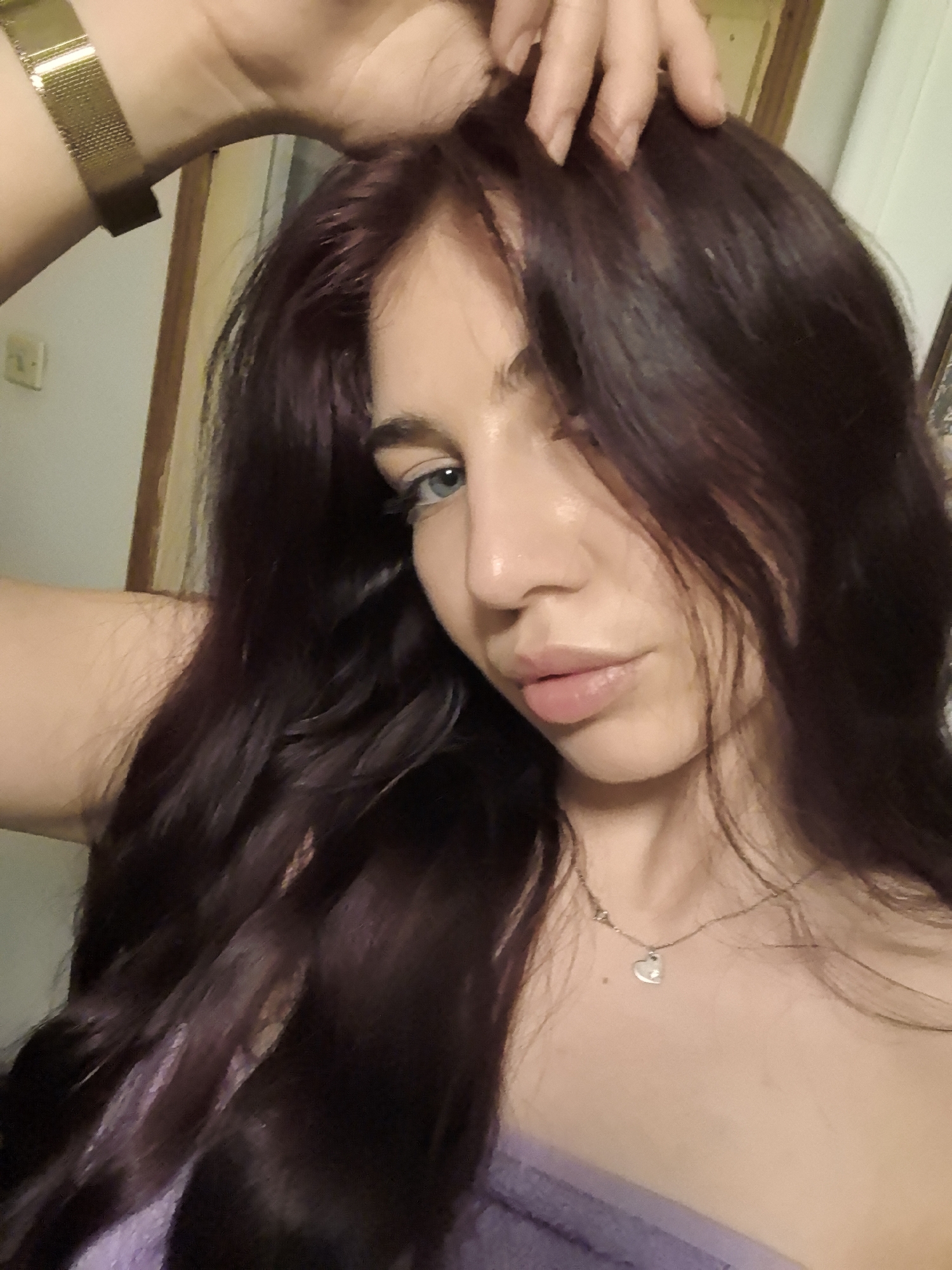 A close-up selfie of a young woman with dark auburn wavy hair touching her head. She has light blue eyes and fair skin. She is wearing a delicate silver necklace with a heart pendant. She also has a gold watch on her left wrist and a purple top. The background includes a light-colored wall and parts of a wooden doorframe. Location: Unknown, Unknown.