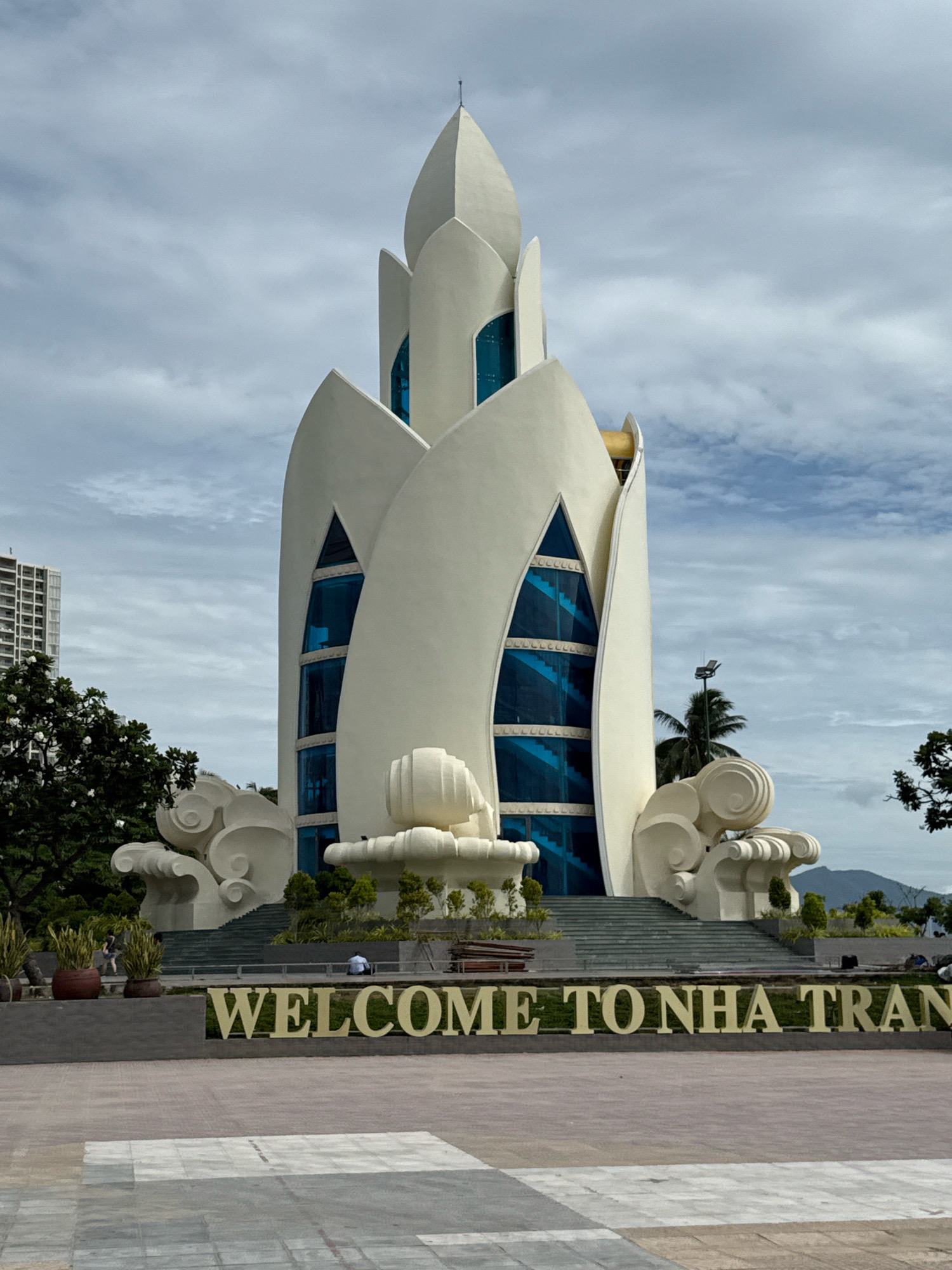 The image shows a monument in Nha Trang, Vietnam, with the words "WELCOME TO NHA TRAN" in large gold letters in the foreground. The monument is a white, petal-shaped structure with blue glass windows and a spire at the top. There are steps leading up to the monument, and decorative cloud-like structures on either side of the base. The environment appears to be a public square or park, with paved walkways, planters with greenery, and trees. In the background, there is a tall building and a cloudy sky. A few people are visible in the image, including one person sitting on a bench and two people walking.