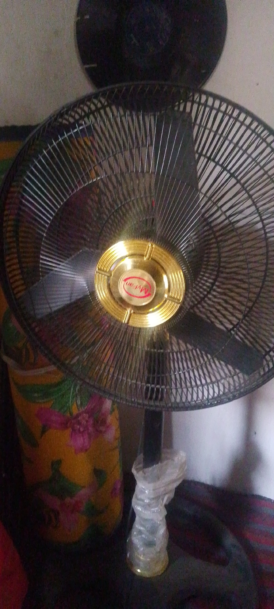 The image shows a close-up view of a black electric oscillating fan. The fan has a protective grille and a circular gold-colored hub with a red logo that appears to read "Myan". The fan is positioned in front of a floral patterned cylindrical object, possibly a container or a fabric-covered item, with yellow and pink flowers. In the background, a dark, circular object, possibly another fan or a wall decoration, is visible. A portion of a white wall is also present. The lower part of the image shows the fan's stand, which is covered in clear plastic. The scene appears to be indoors, and given the context of Jalingo, Nigeria, it likely depicts a domestic or commercial setting where such fans are commonly used for cooling. The lighting suggests it is daytime, but artificial light may also be present.