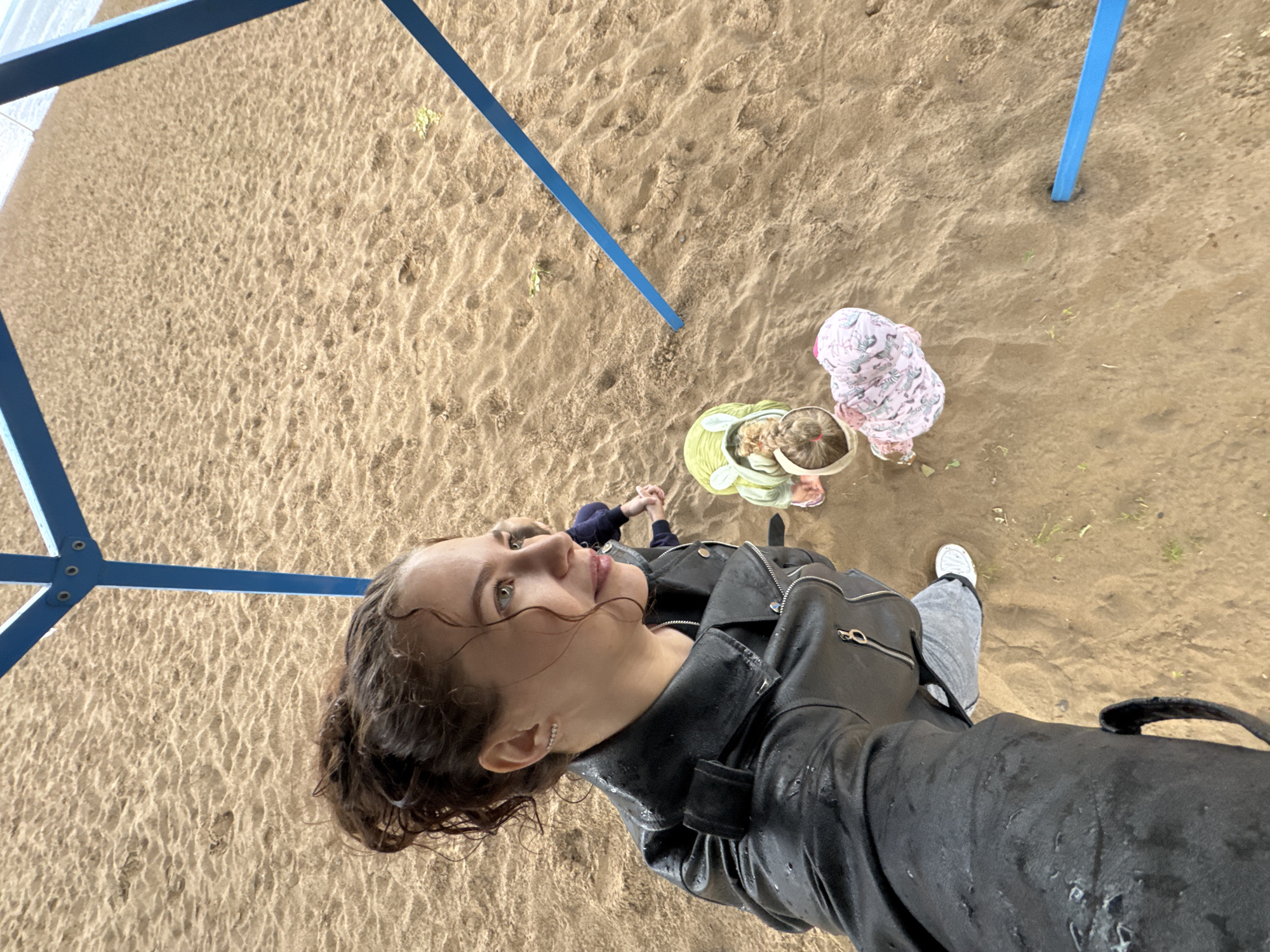 An overhead photograph captures an outdoor scene at what appears to be a sandy playground or beach area in Chaikovsky, Russia. The image is taken from a high angle, with the camera held by a young woman in the foreground. Her dark, curly hair and black leather jacket are visibly wet with rain, suggesting recent or ongoing precipitation. She wears grey pants and white sneakers, and an earring is visible on her left ear. Her face is turned upwards and to the right, with a contemplative expression.
Behind the woman, two young children are visible on the sandy ground. One child, on the left, wears a dark blue top and is holding hands with another person, only partially visible. The second child, closer to the center, is dressed in a light green or yellow top and a hat, looking down. A third child, further to the right, wears a light pink patterned hooded outfit and also appears to be looking down at the sand.
The ground consists of damp, textured sand with subtle ripples and footprints. Several blue metal beams, likely part of a play structure, traverse the frame, reinforcing the playground setting. The scene is illuminated by natural daylight. No visible text is present.