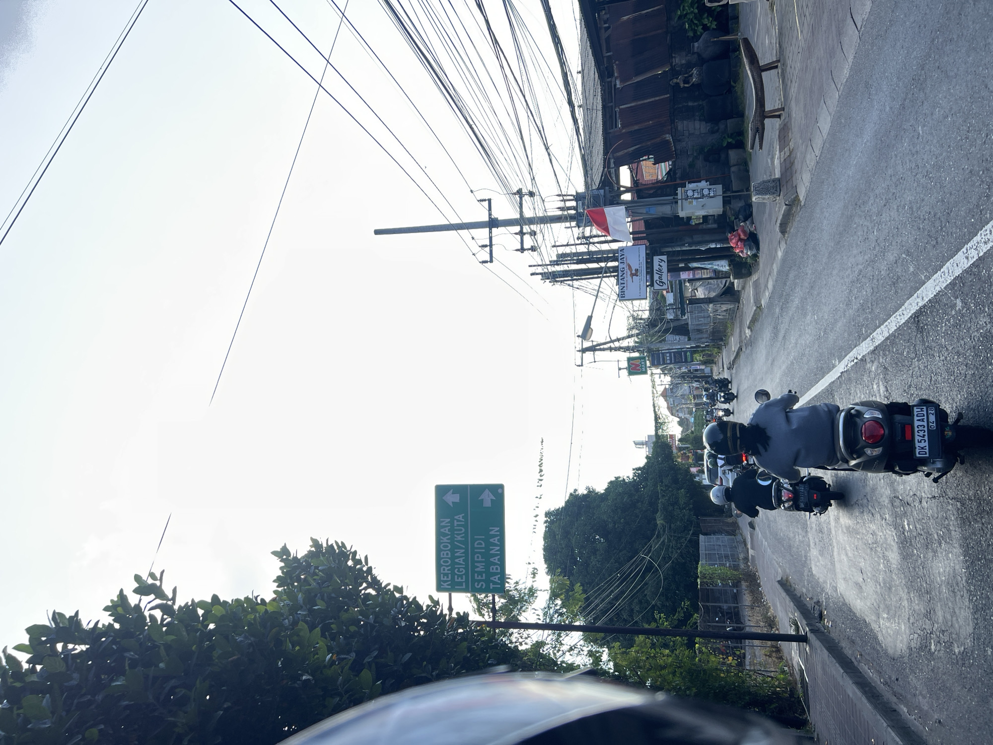 In Denpasar, Indonesia, a road scene unfolds with a focus on transportation and local infrastructure. Several individuals on motorcycles navigate the asphalt road, suggesting a common mode of transit. A green directional signpost indicates routes to "KEROBOKAN LEGIAN/KUTA" and "SEMPIDI TABANAN," providing geographical context. Power lines crisscross the sky, a ubiquitous element in many urban and suburban landscapes, indicating a developed electrical grid. On the right side of the road, buildings with weathered metal roofs line the street, alongside a bench and what appears to be decorative statues. The overall environment suggests a bustling street in a tropical climate.