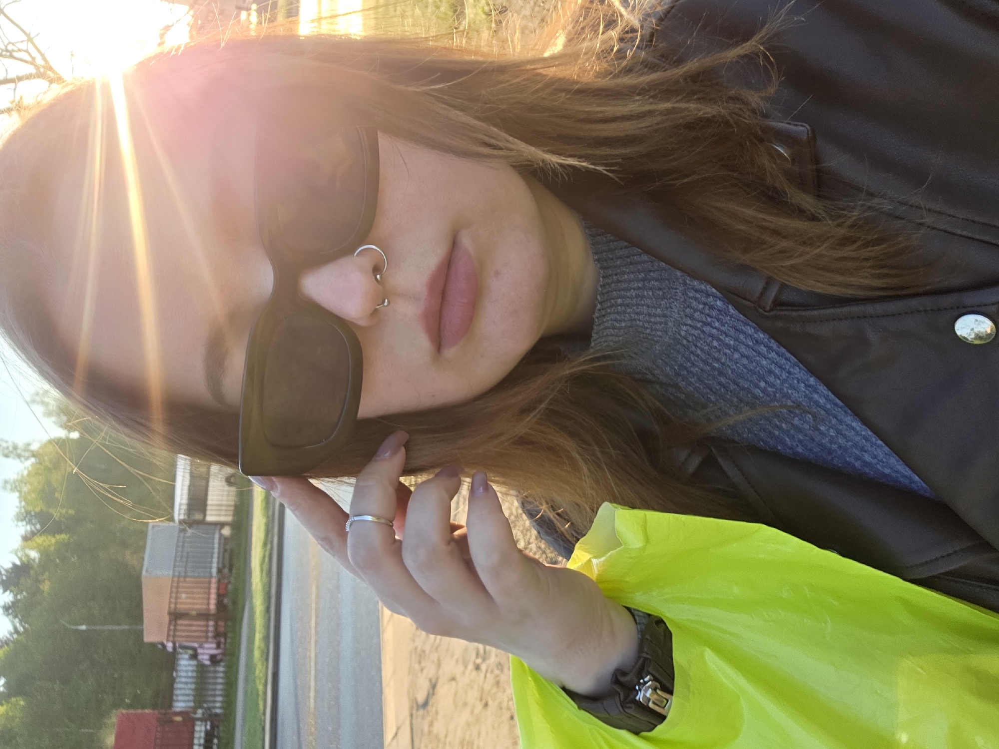 A woman with light brown hair and sunglasses is the central focus of this outdoor photograph. She has a nose ring and is wearing a gray sweater under a dark jacket. Her left hand, adorned with a ring, is visible. A bright yellow bag is also partially visible. In the background, there are trees and some buildings that appear to be storage containers or industrial structures, alluding to a location possibly near a transportation hub. The sun's glare is prominent in the upper left corner, obscuring part of the scene.