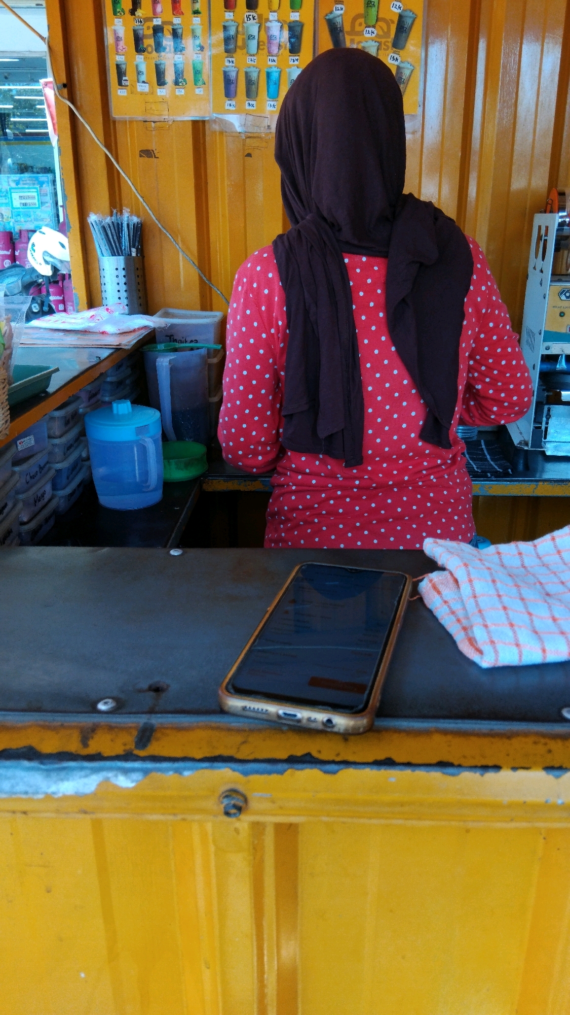In the city of Watesalit, Indonesia, a person wearing a red and white polka dot shirt and a dark brown hijab stands behind a counter, facing away from the camera. They are positioned in what appears to be a food or drink stall. In front of the person, on the counter, is a smartphone with a gold-colored case. To the right of the phone, a checkered cloth with orange and white squares is placed. The stall is decorated with a yellow corrugated metal wall. Various items, including cups, containers, and a blue water jug, are visible on shelves and the counter. Above the stall, a menu board displays images of drinks. The overall scene suggests a commercial setting, likely a small business operating outdoors or in a covered open-air space.