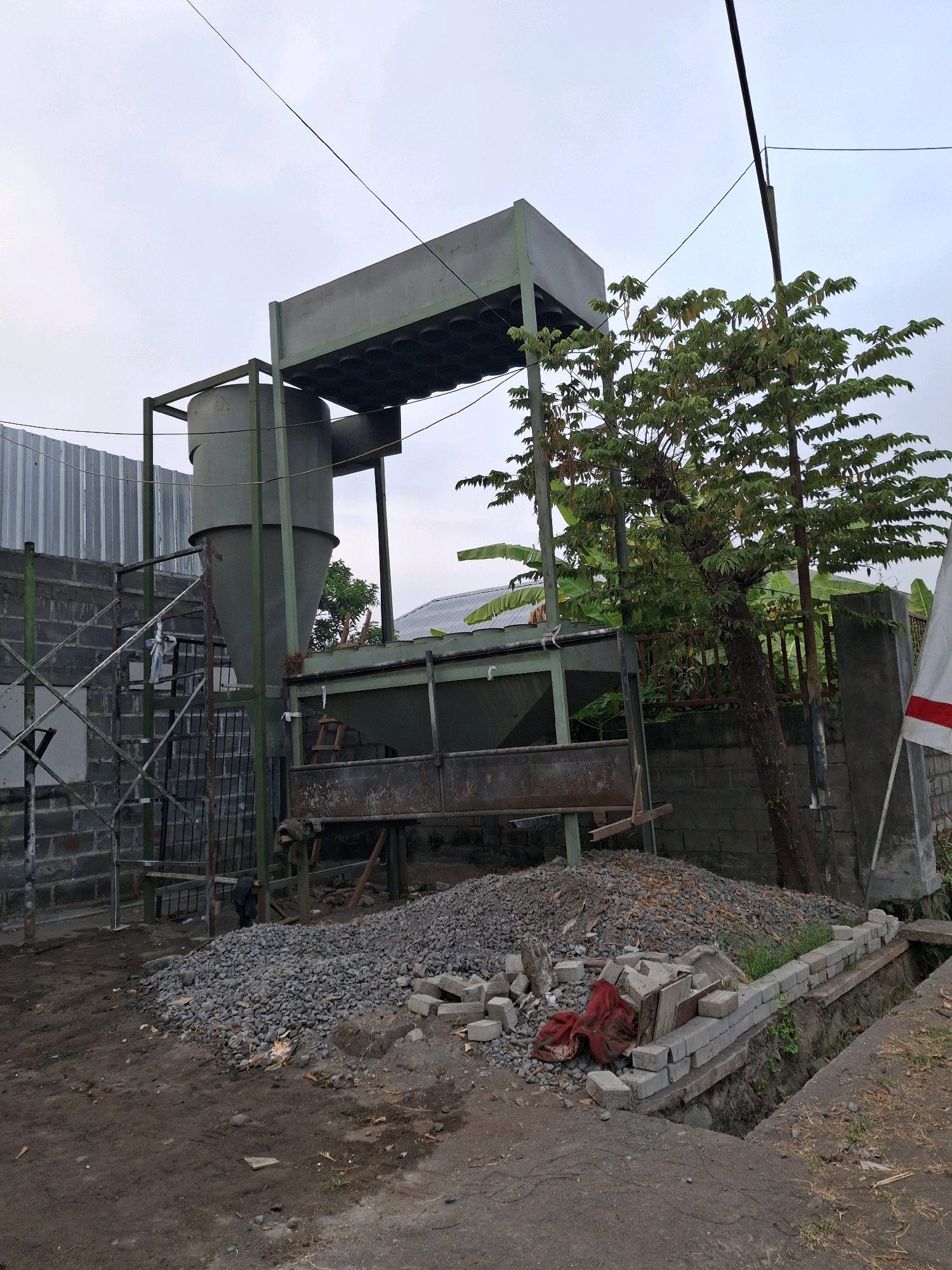 An outdoor industrial site in Lumajang, Indonesia, is depicted. The scene is dominated by a large, gray, conical silo situated within a metal frame structure. Adjacent to the silo, a rectangular elevated platform with a grid of circular openings is visible, supported by a metal frame. A metal staircase leads up to a platform near the silo. The structure appears to be part of a processing or storage facility. In the foreground, a pile of gravel and some loose bricks are scattered on the dirt ground. A tree with green foliage grows to the right of the silo, near a brick wall. A white flag with a red stripe is visible on a pole to the far right. The environment is overcast, suggesting cloudy weather.