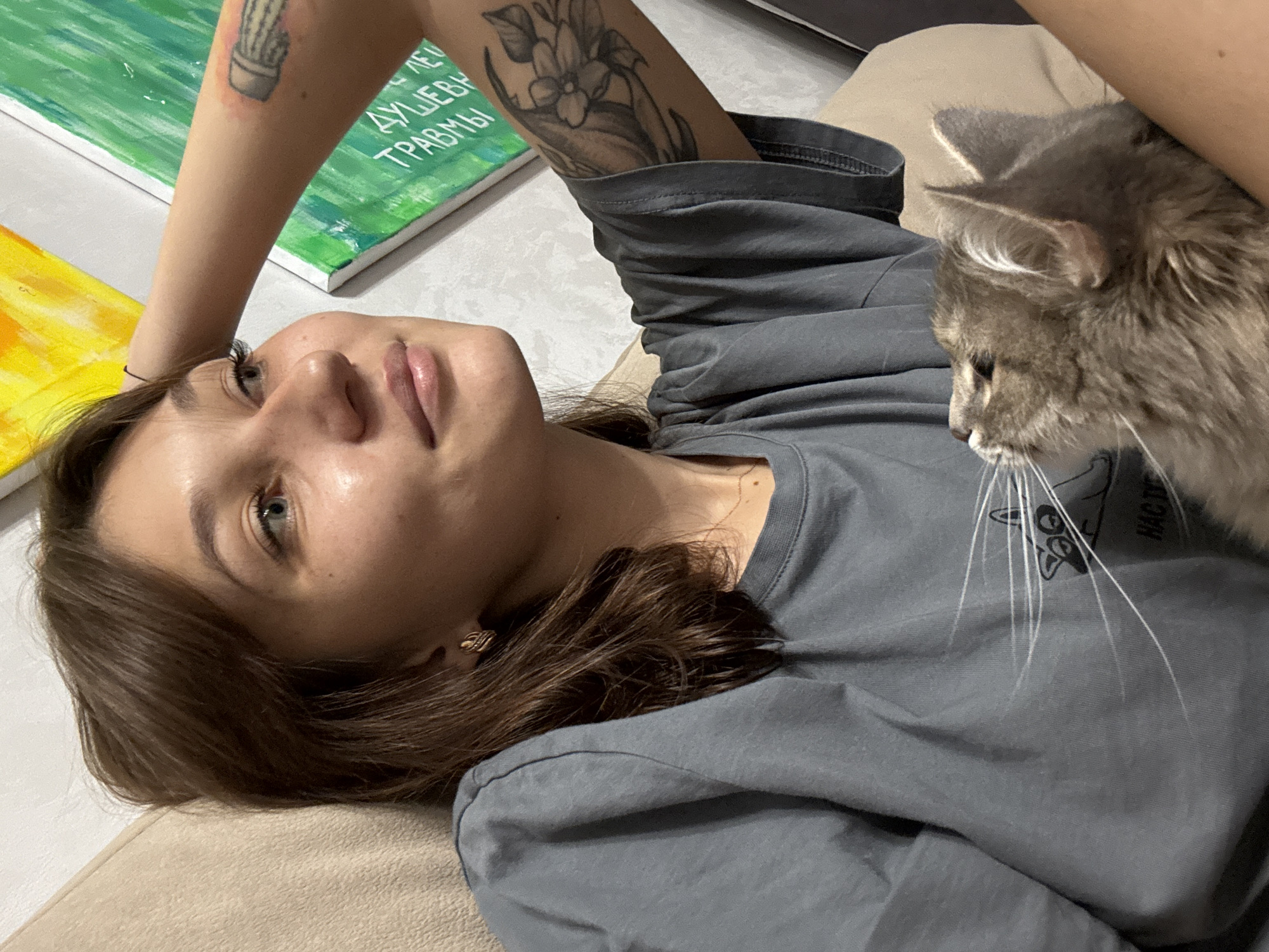 A young woman with light brown hair and blue eyes is shown from the chest up, lying on a beige sofa. She is wearing a grey t-shirt and has her left arm raised behind her head. Her right arm is holding a grey cat. She has tattoos on her right arm, one of which appears to be a flower. Behind her, on the wall, are two paintings. One is abstract with yellow and orange colors, and the other is green with white text. There is a faint pattern on the wall, and the texture of the sofa is visible. The lighting is soft and appears to be natural. The image is taken indoors, likely in a residential setting. There are no discernible landmarks or details that would suggest a specific location within Novokuznetsk, Russia.