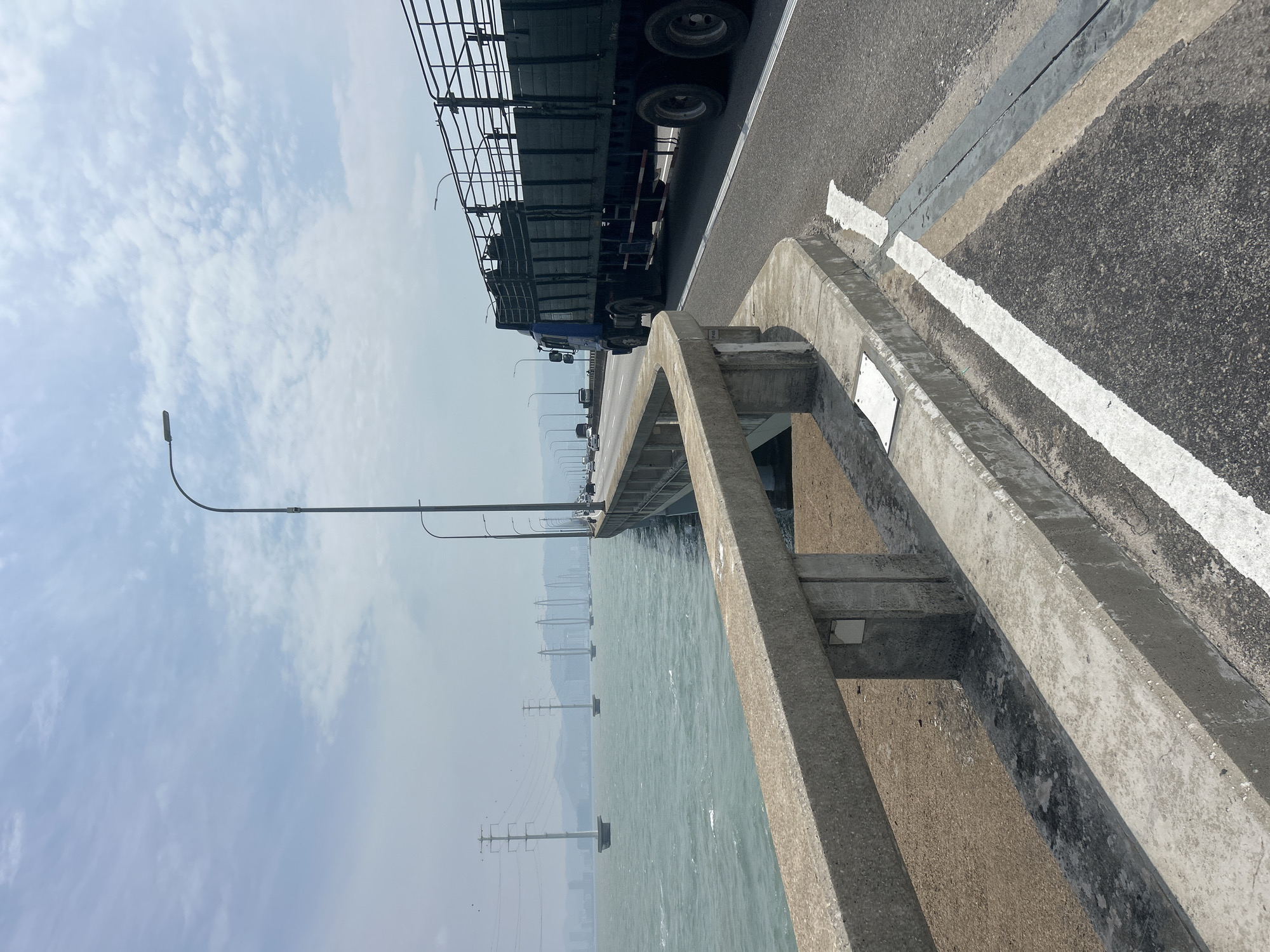 A truck travels down a multi-lane highway on a bridge in George Town, Malaysia. The truck is on the right side of the road. The highway is lined with streetlights and a concrete barrier separating the roadway from a body of water on the left. The water is choppy, with whitecaps visible. In the distance, several large, pylons with power lines stretch across the water. The sky is overcast with scattered clouds.