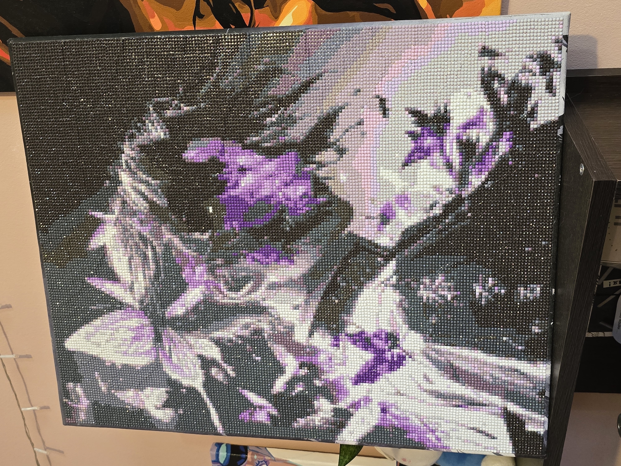 A framed diamond painting depicting a close-up of a woman's face with flowing, light-colored hair is displayed on a shelf. The painting uses a palette of purples, pinks, whites, and grays against a dark background. In the background, a string of small, clear lights hangs across a light pink wall. To the right of the diamond painting, another framed artwork with abstract orange and black shapes is partially visible. The shelf is dark wood, and below it, a portion of what appears to be a book or magazine is visible. The lighting suggests an indoor setting, possibly a room or studio. There are no discernible geographic markers to indicate a specific city or location.
