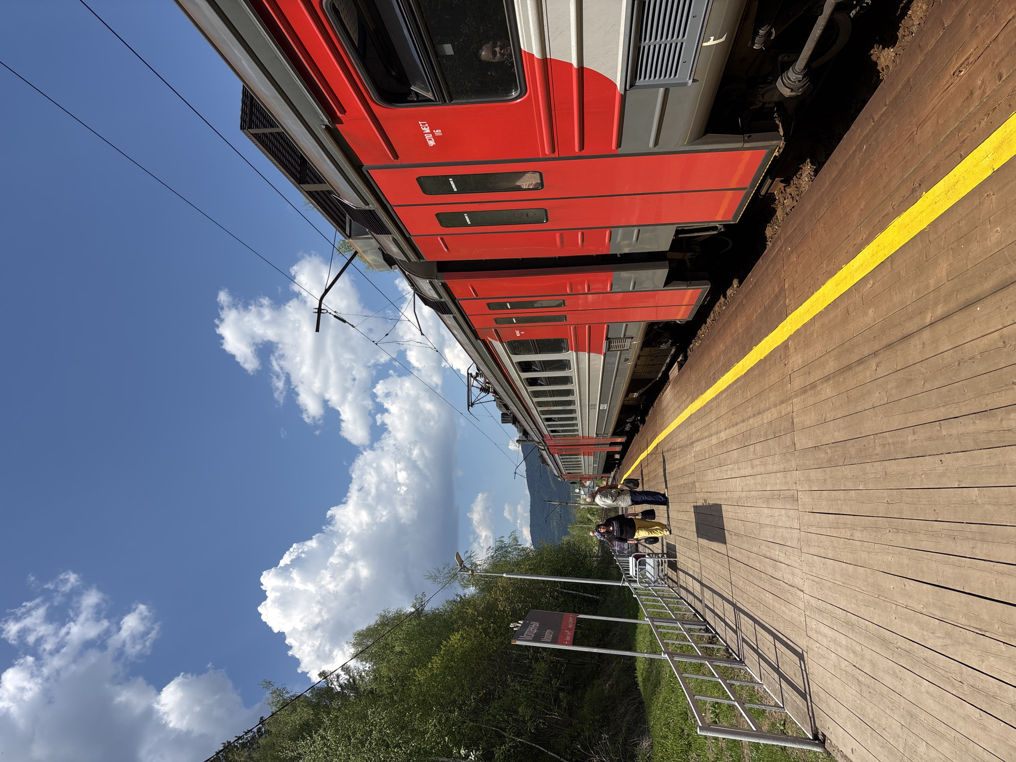 Scene: A bright, sunny day at the train station in Овсянка, Russia.
Subjects: A red and gray passenger train stands adjacent to a wooden platform, with a yellow safety line painted along its edge. Two people stand on the platform beside a railing.
Objects: The train, platform, sky, clouds, trees, mountain in the background, railing, sign.
Actions: People standing and waiting on the platform. The train is stationary.
Interactions: People waiting for the train.
Environment: The train station is bordered by trees and greenery, with a view of mountains in the distance. The sky is blue with scattered white clouds.