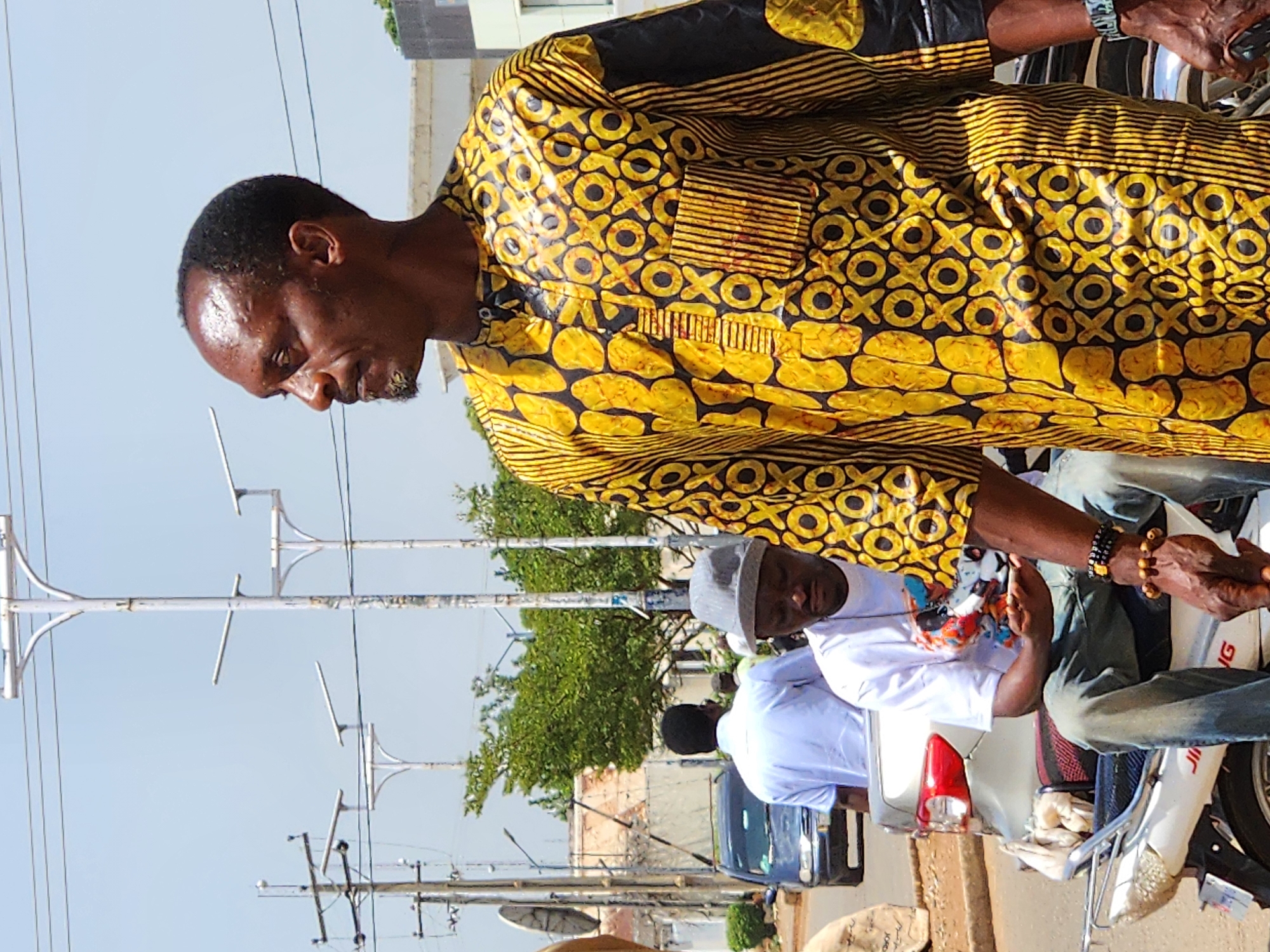 In an outdoor urban scene, likely on a street in Jos, Nigeria, on a bright, clear day with a pale blue sky, several individuals are visible. The most prominent figure is an adult male, facing to the right in profile. He has dark skin, short dark hair, and a light beard. He is wearing a vibrant yellow and black patterned garment with a short-sleeved, collared top. The pattern features black circles and 'X' shapes, interspersed with irregular yellow shapes and vertical black lines. On his right wrist, he wears a dark watch.
In the middle ground and slightly to the left, another man is seated on a white motorcycle. He is wearing a white t-shirt with a colorful graphic design and blue jeans. He has a grey cap on his head and is looking downwards, holding a mobile phone in his right hand. His left arm rests on his lap, adorned with bracelets. The motorcycle is white with silver accents and a visible license plate.
Further in the background, another man stands wearing a white short-sleeved shirt and dark trousers. He is facing away from the camera. A dark blue car is parked on the side of the road behind him, with trees and buildings forming the backdrop of the environment. Streetlights and power lines are visible in the sky. The overall impression is of a bustling street scene in a Nigerian city.