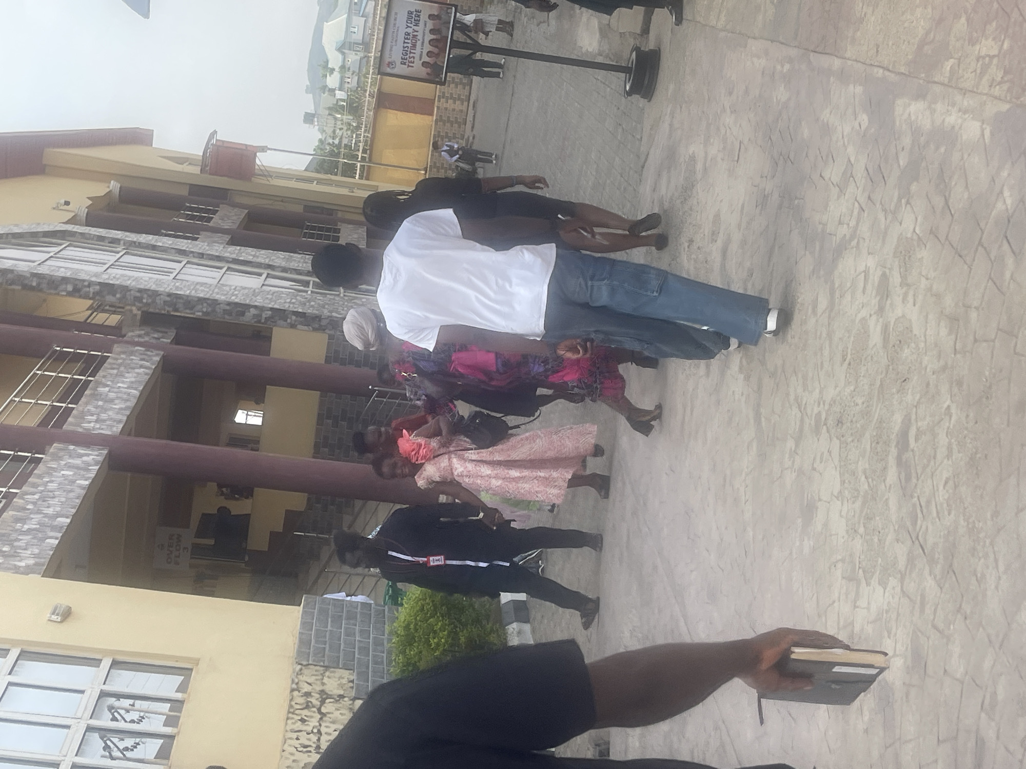 The image shows a group of people standing on a paved area outside a multi-story building. A sign that reads "LIVING FAITH CHURCH" and "REGISTER YOUR TESTIMONY HERE" is visible. The building has a light yellow facade with windows and some columns. The location appears to be in Abuja, Nigeria. One person is holding a notebook.