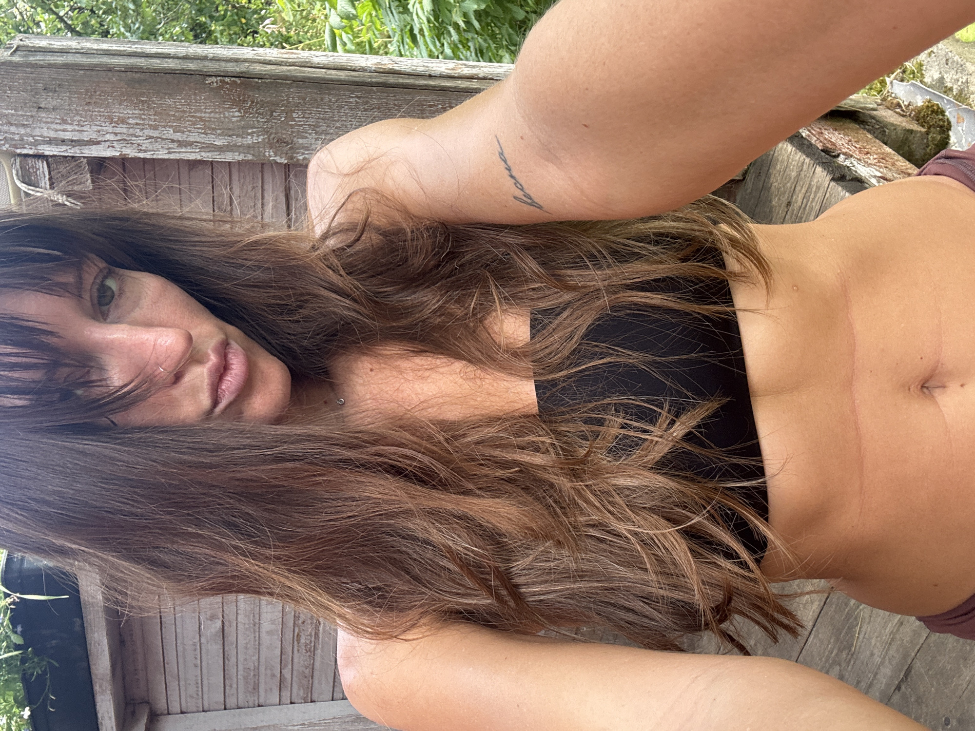 A medium shot captures a young adult with long, dark brown hair, bangs obscuring part of her face, and a slight pout. She is wearing a black bandeau top and appears to be outdoors in a natural setting, possibly a garden or backyard. The background consists of weathered wooden structures and lush green foliage. Her left arm is raised, and a small tattoo is visible on her forearm. The lighting suggests it is daytime. The image is a selfie, indicated by the angle and framing. The location is identified as Krasnaya Presnya SNT, Russia.