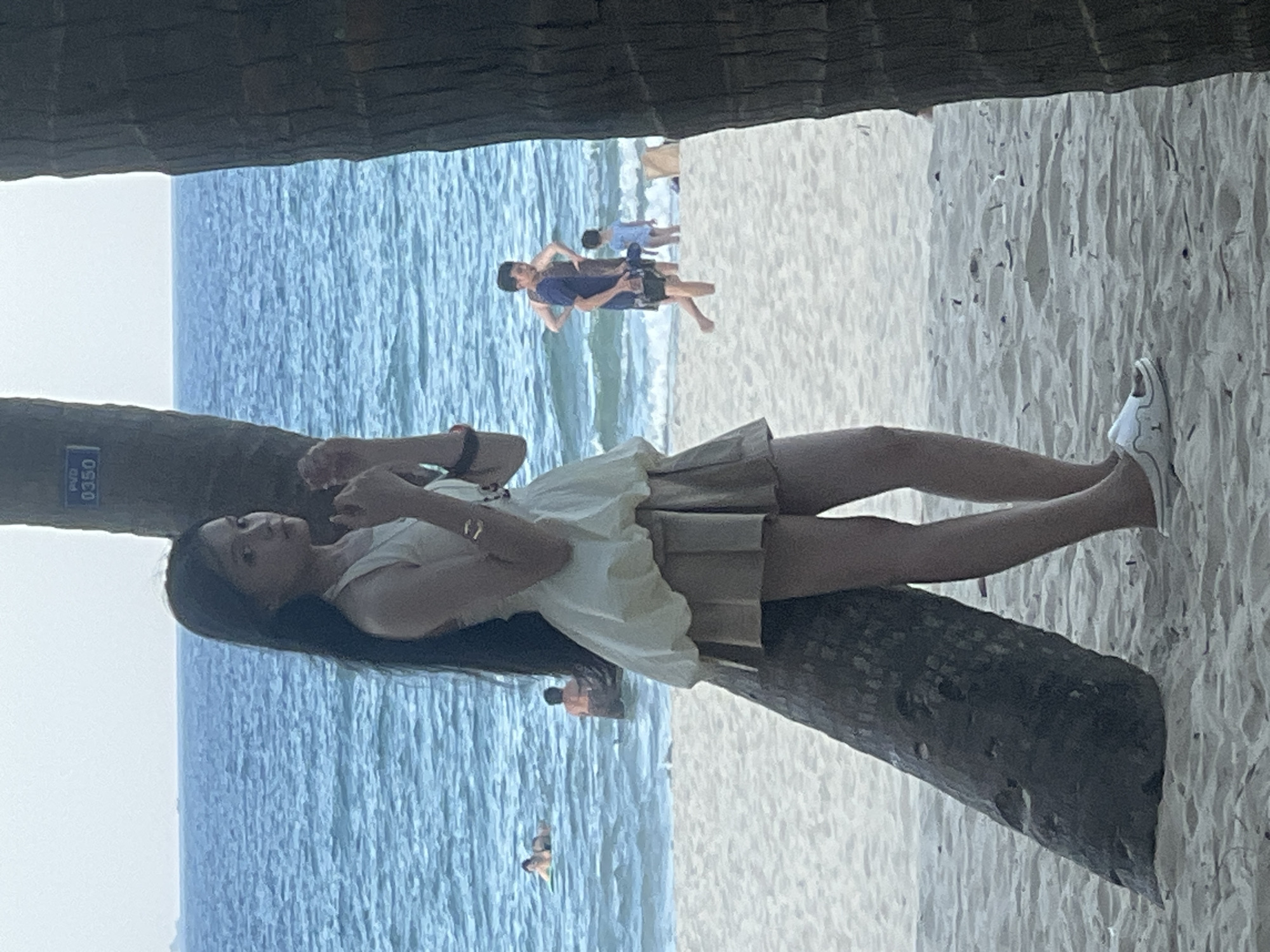 This image depicts a beach scene in Đà Nẵng, Vietnam. The primary subject is a woman lying on what appears to be a fallen palm tree trunk, with her legs extended towards a sandy beach. She is wearing a light-colored, possibly cream or white, dress and white sneakers. Her hair is long and dark.
In the background, the ocean is visible with gentle waves. Several other people are present in the water, appearing to swim or wade. Further back on the beach, two individuals are walking, one in a blue shirt and shorts, and another in a lighter colored shirt. The scene is set under a canopy, possibly formed by palm trees, which creates a dark frame at the top and sides of the image.
The lighting suggests it is daytime, likely late afternoon or early evening, given the softer light and potential shadows. The weather appears pleasant for a beach outing.
A small blue and white rectangular sign is attached to the palm tree trunk in the foreground, bearing the text "PVD 0350". The overall mood is relaxed and leisurely, characteristic of a beach vacation.