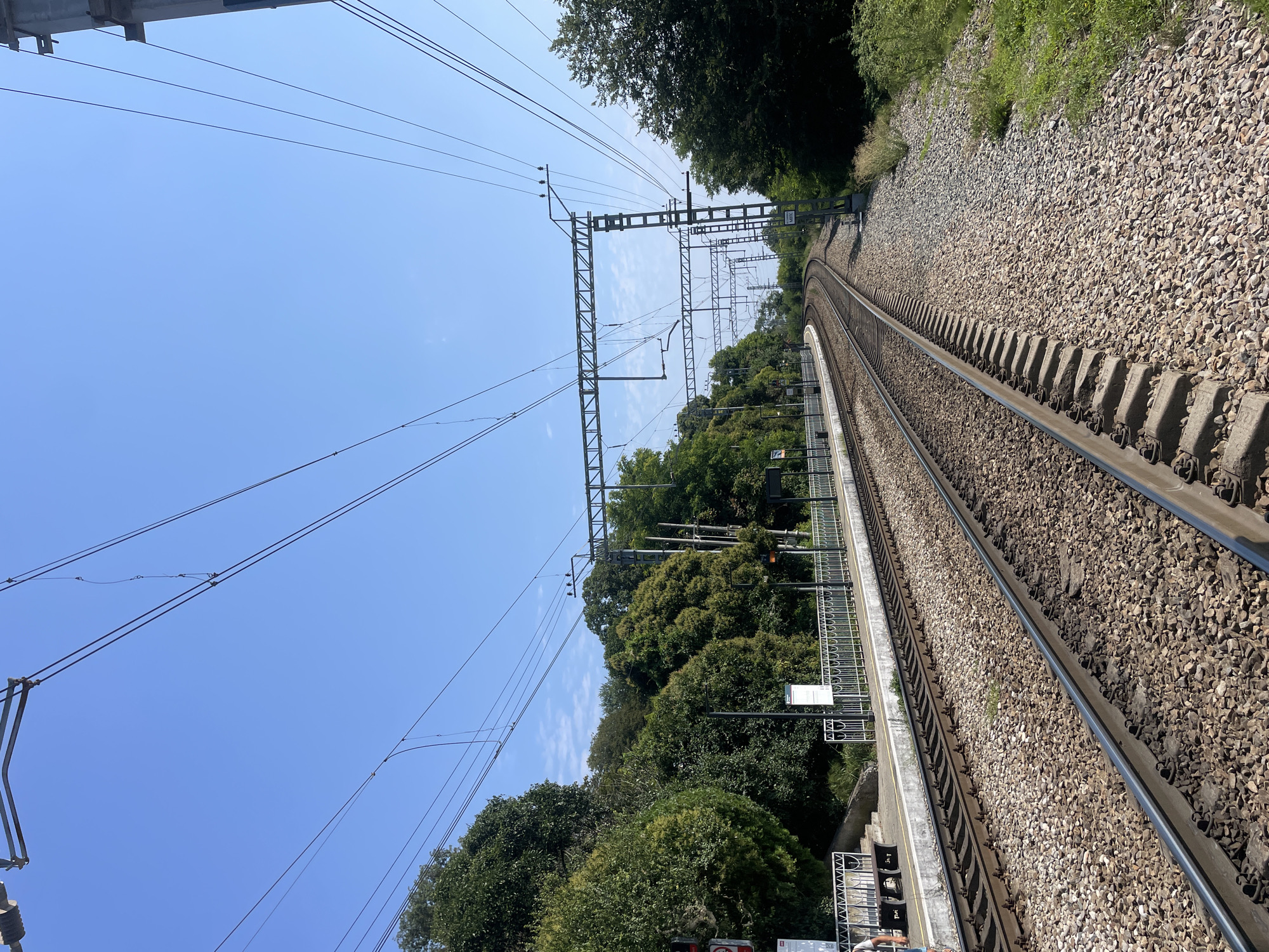 The image shows a view of train tracks and a platform at a train station. The tracks are surrounded by ballast and have overhead electrical wires. There are trees and vegetation on either side of the tracks and a clear blue sky above. The platform has a fence, benches, and signs. The scene is likely in Sochi, Russia, as suggested by the architectural style and vegetation. There are no people visible in the immediate foreground or interacting with the environment. The overall impression is one of a quiet, functional train station on a sunny day.