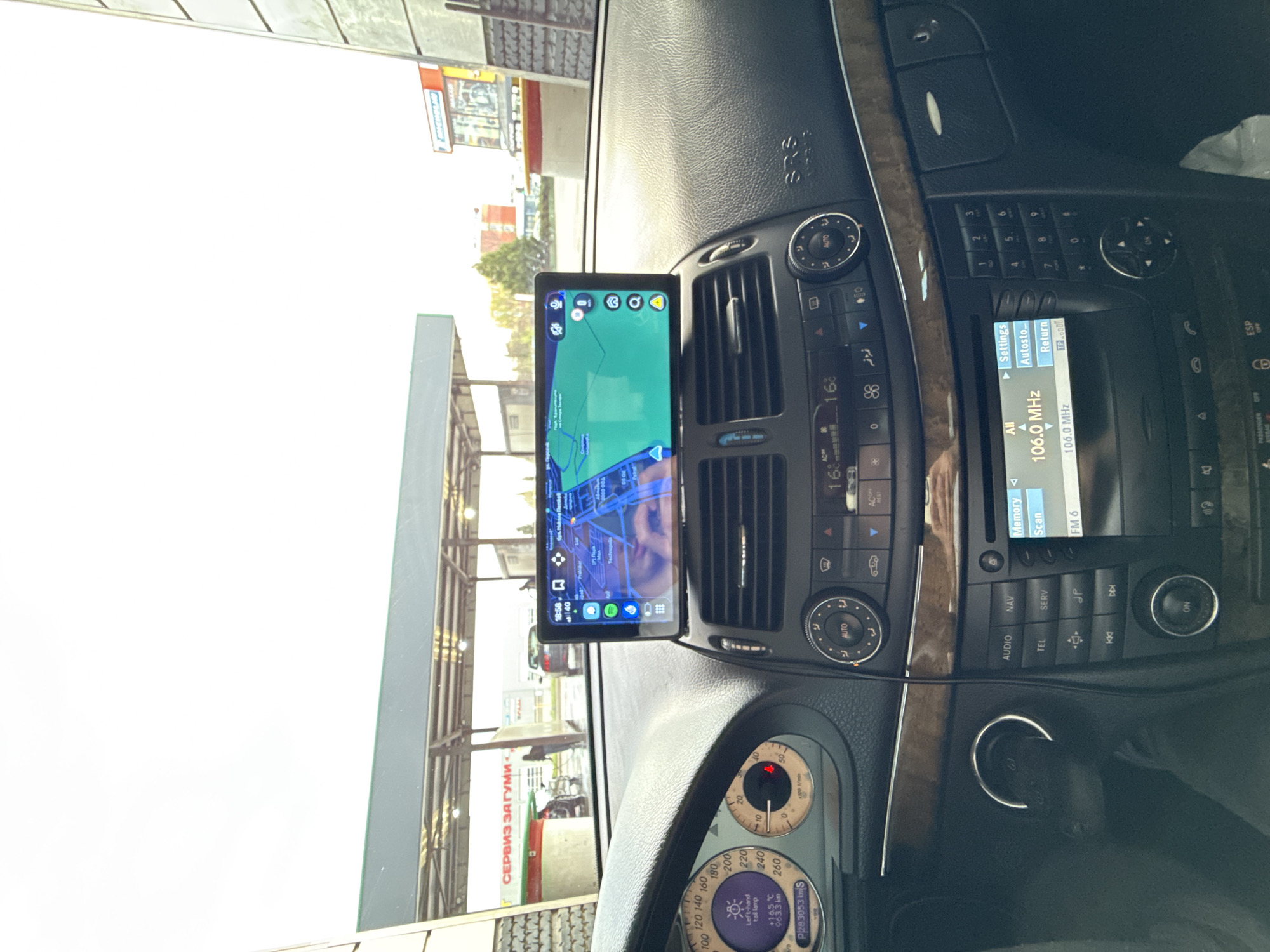 The image shows the interior of a car, focusing on the dashboard and the driver's perspective looking out the windshield. A GPS navigation system is mounted on the dashboard, displaying a map. Outside the car, visible through the windshield, is a car service station identified as "СЕРВИЗ ЗА ГУМИ" (Tire Service) written in Cyrillic script, indicating that the location is in a Cyrillic-using region. Based on the language, it's plausible the location is in Stara Zagora, Bulgaria. The car's instrument panel is visible, along with the radio and climate control system.