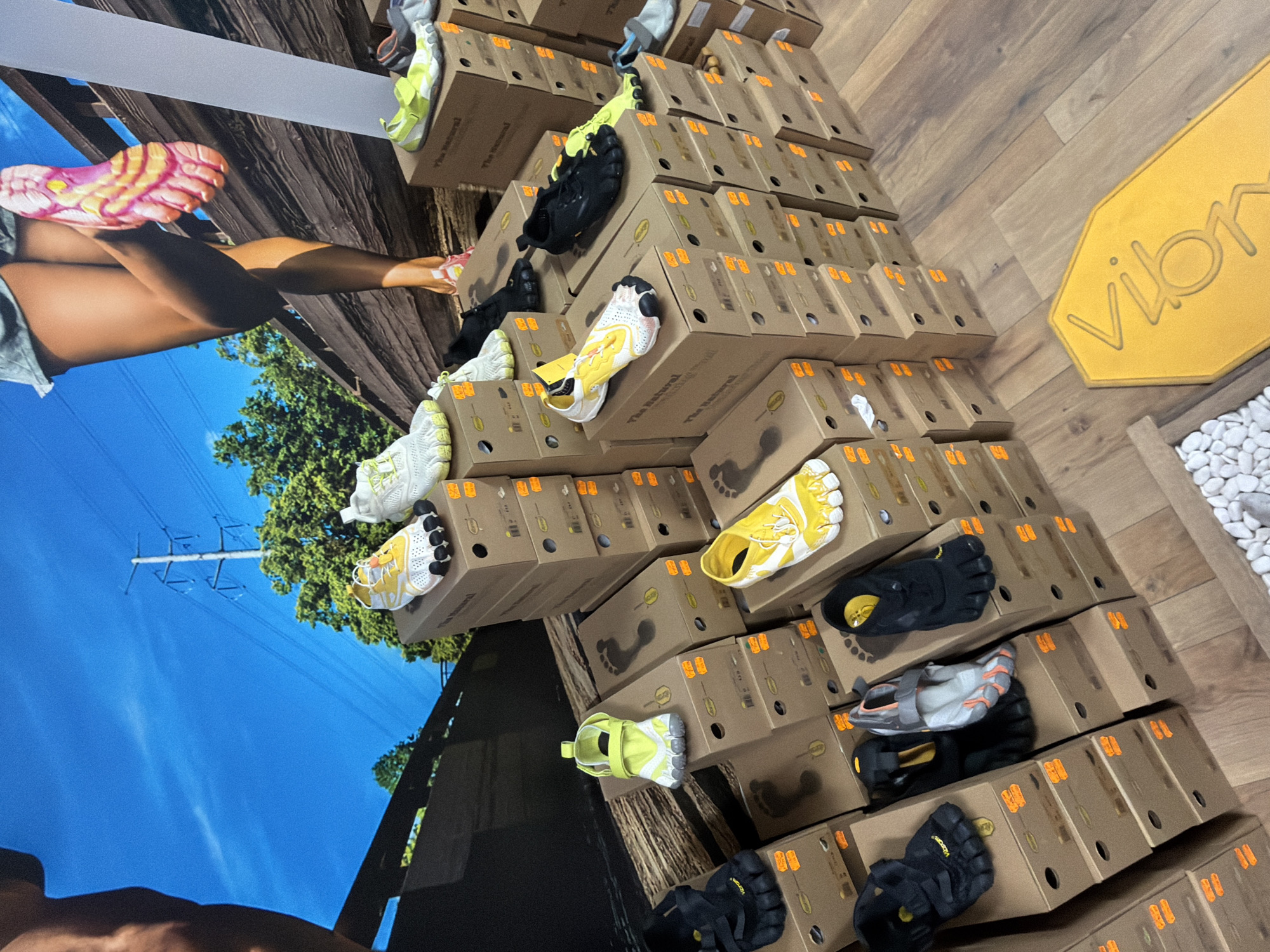 The image depicts a display of Vibram FiveFingers shoes in what appears to be a retail environment. Stacks of cardboard boxes, each containing a pair of shoes, dominate the scene. The shoes are visible atop the boxes, showcasing various styles and colors. A yellow Vibram logo is visible on the right side, near a decorative arrangement of white pebbles. A large promotional graphic is on the left wall showing a woman's legs and feet wearing Vibram FiveFingers, in front of an outdoor background of trees and a blue sky. There are no visible individuals. The scene seems to be indoors, with wood-grained flooring, likely inside a shoe store. Location information about Sofia, Bulgaria, is not evident from the image.