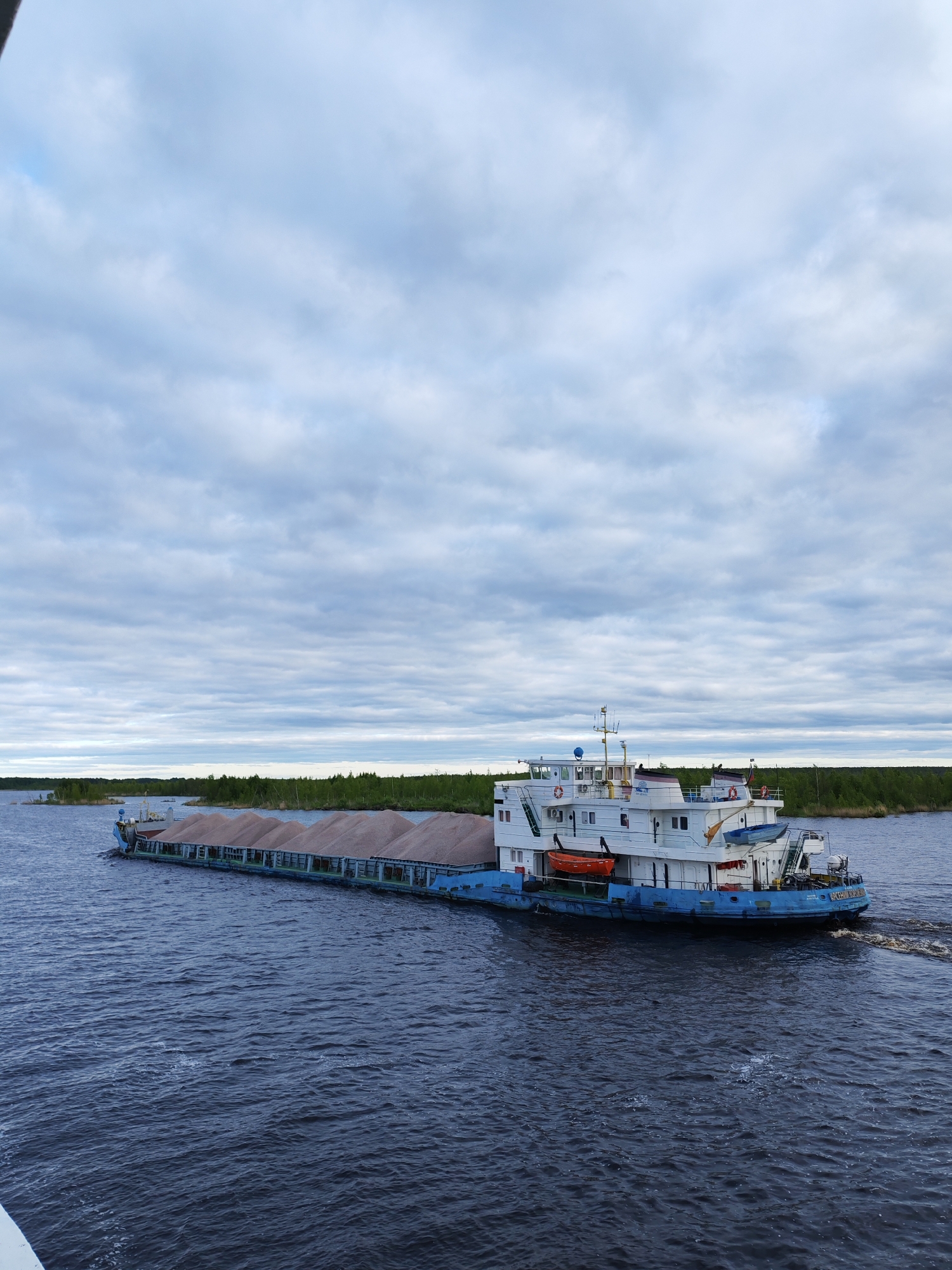A long cargo barge navigates a wide, dark waterway under a cloudy sky. The barge is laden with a mound of brownish material, presumably gravel or sand, and is marked with “ЯРОСЛАВ МУДРЫЙ” on its lower hull. The barge's main structure is white with a blue lower hull and features multiple windows, life preservers, and a small crane on the top deck. The water surface is rippled, indicating movement. A distant shoreline with green vegetation is visible in the background. The barge's position and the presence of vegetation along the shoreline suggest a setting near a river or lake. It is unknown if this is near the city of Bereg, Russia.