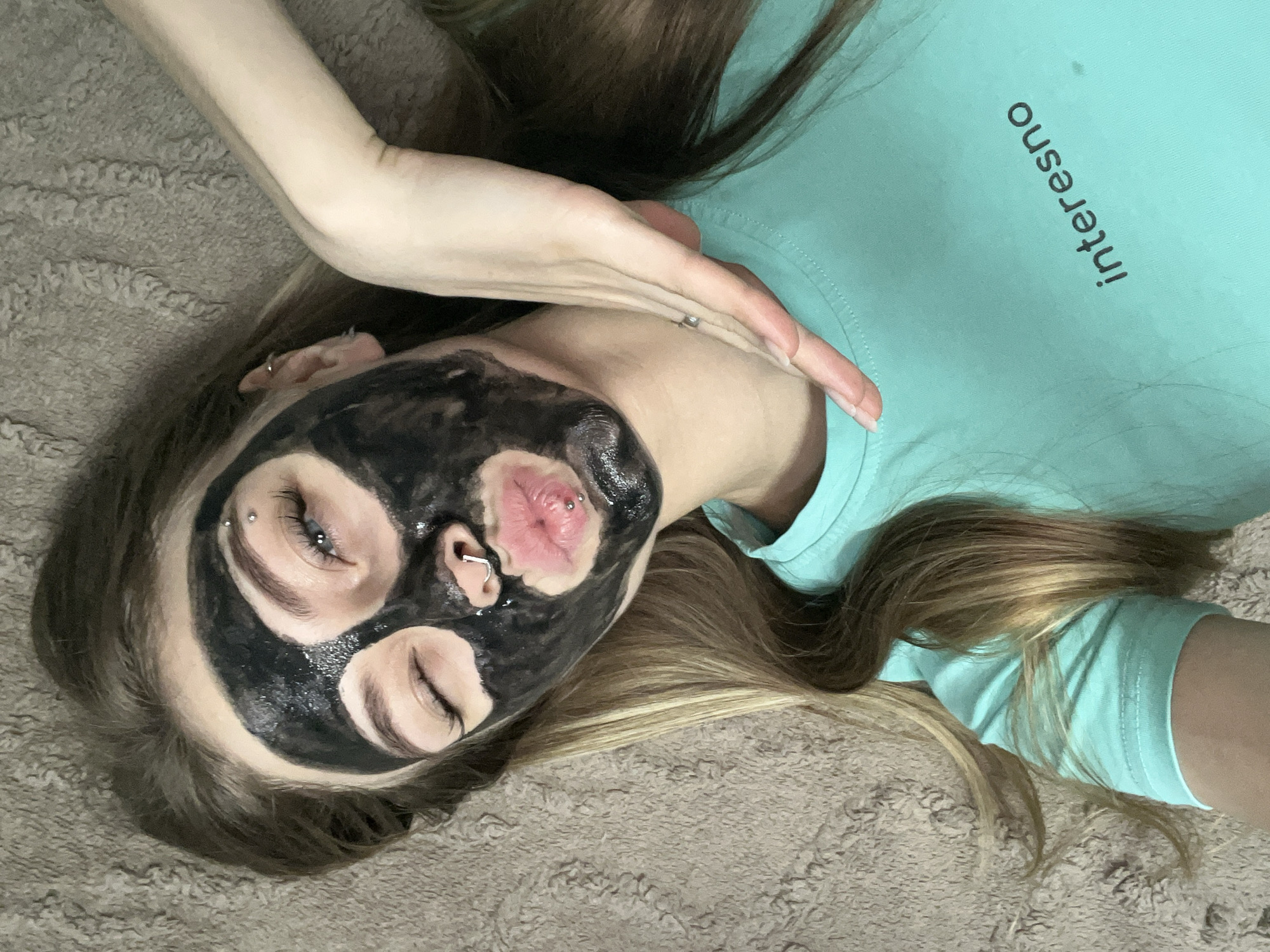 A woman is lying on a textured, neutral-colored surface, possibly a rug or blanket. She is wearing a light teal t-shirt with the word "interesno" printed on it. The woman has a black facial mask applied. She has piercings in her eyebrow, nose, and lip. Her hair is blonde with darker roots. Her right arm is raised, with her hand near her neck. The location is not discernible and there is no evidence to indicate Фоки, Russia.