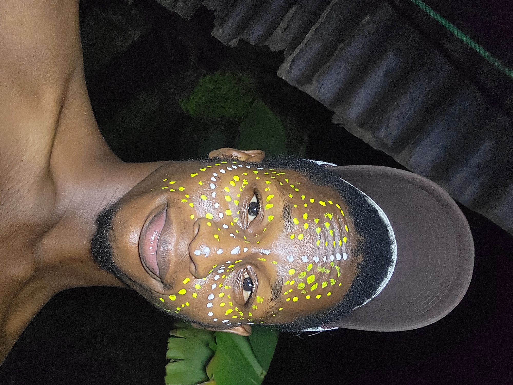A close-up, tilted photograph depicts a Black male, likely in his late teens to early thirties, wearing a brown baseball cap and adorned with yellow and white paint dots on his face. His skin is light brown, and he has a short black beard. The man is facing the camera with a slight smile, his mouth open. The background is dark, but green leaves are visible near the bottom, and corrugated metal sheets appear in the upper right. The scene likely takes place outdoors at night, potentially in a residential or semi-urban area, possibly Nkpor, Nigeria, based on contextual information. The lighting is poor, with the subject appearing illuminated by an unknown source.