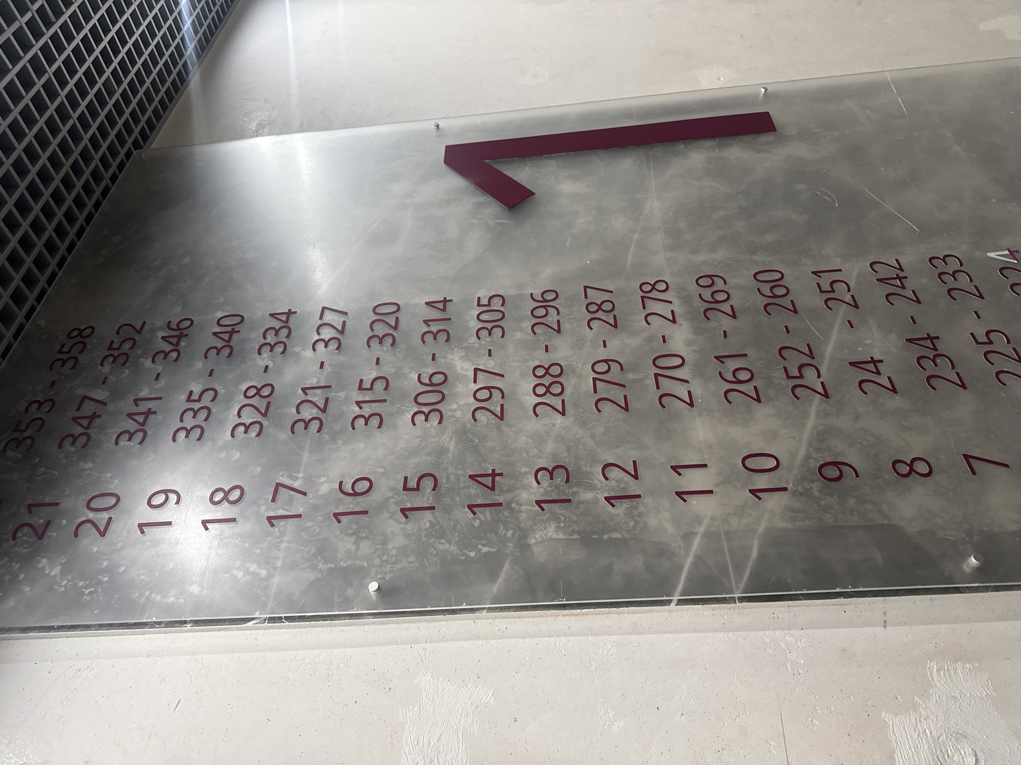 A directional sign, indicating room numbers 225-358 in Saint Petersburg, Russia, is displayed on a clear, flat surface, likely made of glass or acrylic. The room numbers, grouped in ranges (e.g., 353-358 for room 21), are printed in a reddish-purple hue along with corresponding numbers 7-21. A large reddish-purple arrow points to the left, indicating the direction of the rooms. The sign is positioned near a wall with a grid-like pattern. The overall environment appears to be a hallway or corridor within a building.