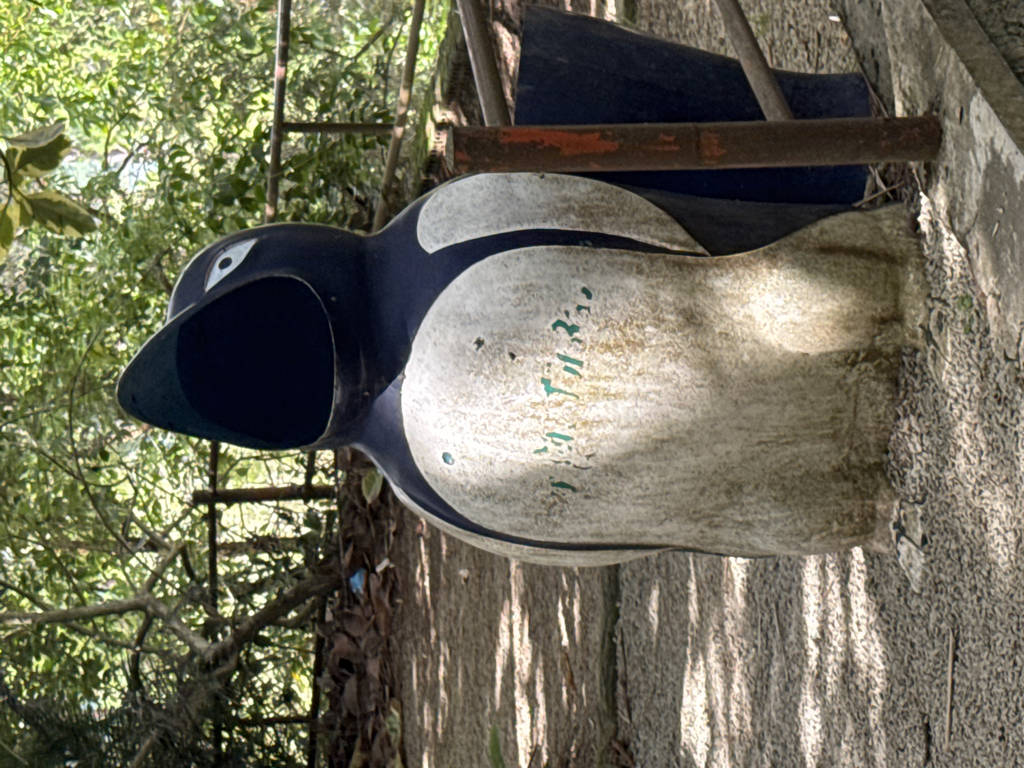 A life-sized, open-mouthed penguin statue, likely serving as a trash receptacle, is situated outdoors in a gravel-covered area. The statue is predominantly black and white, with some green markings on its white chest. It is positioned in front of lush green foliage and trees, with a railing visible in the background. The surrounding environment suggests a park or a similar public outdoor space in Da Lat, Vietnam. The lighting indicates it is daytime, with shadows cast by the trees and sunlight dappling the ground. There are no people visible in the image.