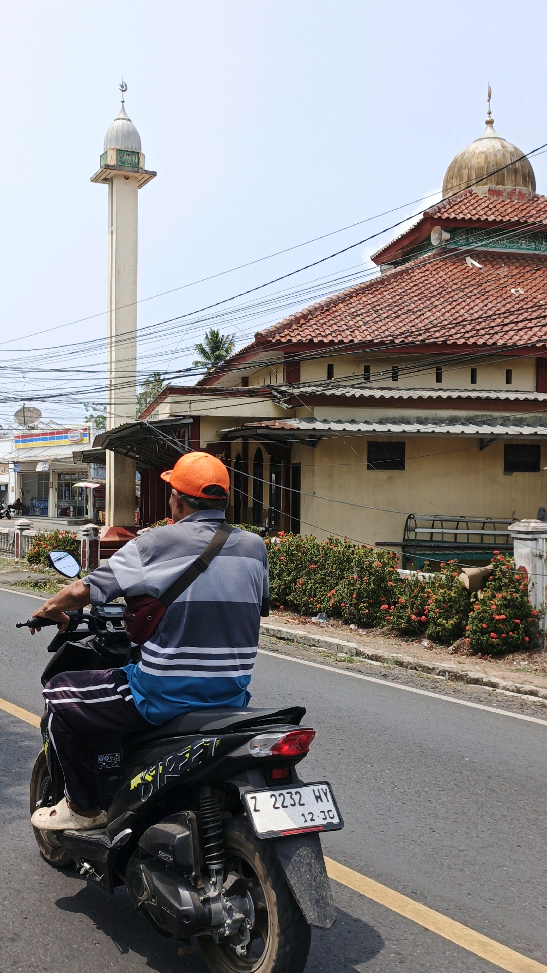A man wearing an orange cap and a gray and blue striped shirt rides a black scooter on a paved road. He is facing away from the camera, looking towards a building with a red tiled roof and a golden dome. To the left of the building is a tall, slender minaret with a crescent moon on top. The building appears to be a mosque. The scene is set in Desa Wonoharjo, Indonesia, with lush green bushes with red flowers lining the road. Power lines crisscross the sky. In the background, a row of shops is visible, one of which appears to be an Indomaret. The sky is clear and bright.