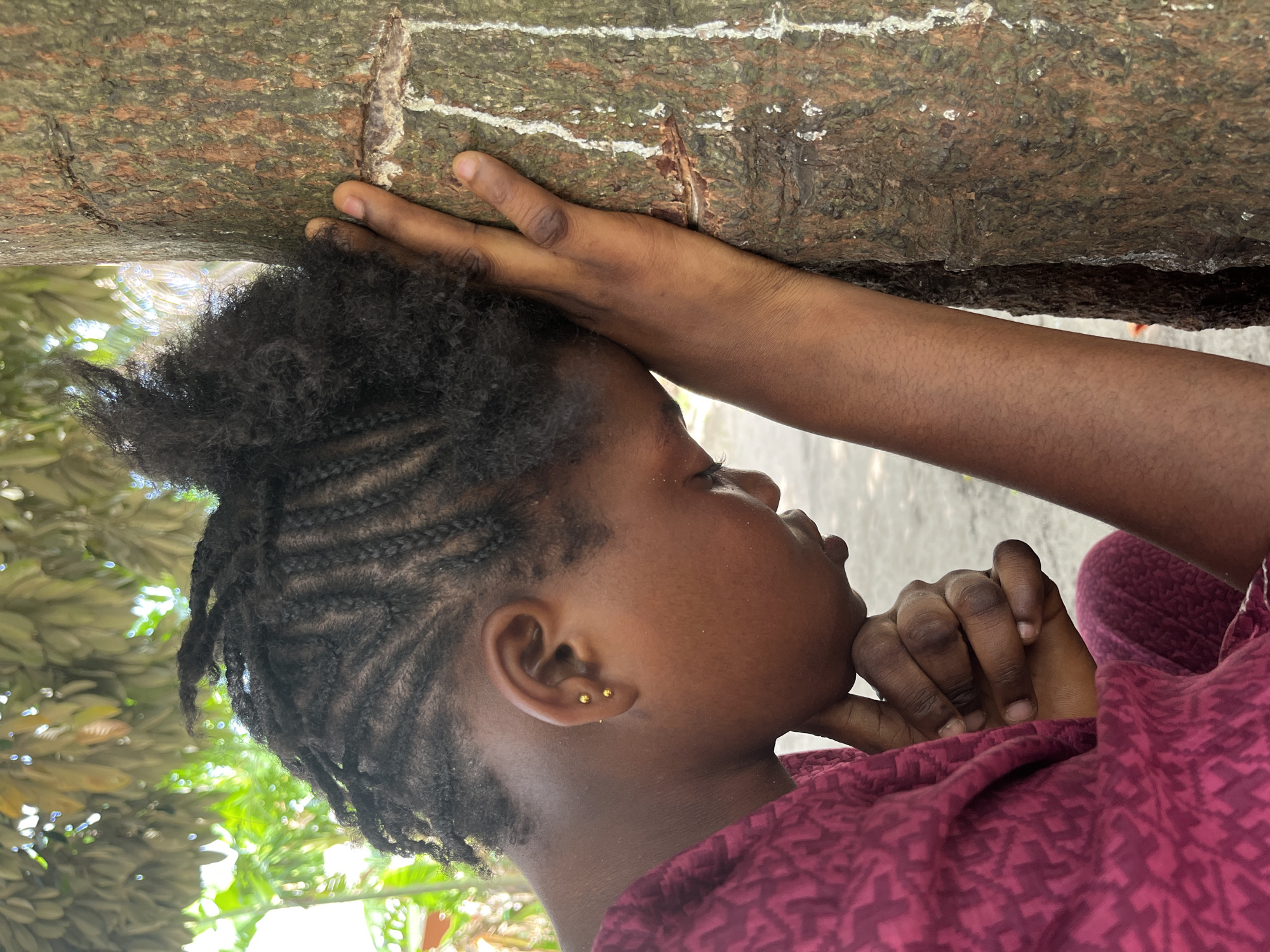 A young person with braided hair and two earrings is leaning against a tree. The person's left arm is extended upward, and their hand is pressed against the tree trunk. The person's right hand is held near their face, with fingers touching their chin. The person is wearing a maroon shirt. The image was taken outside, with foliage visible in the background. The location is not mentioned in the image.