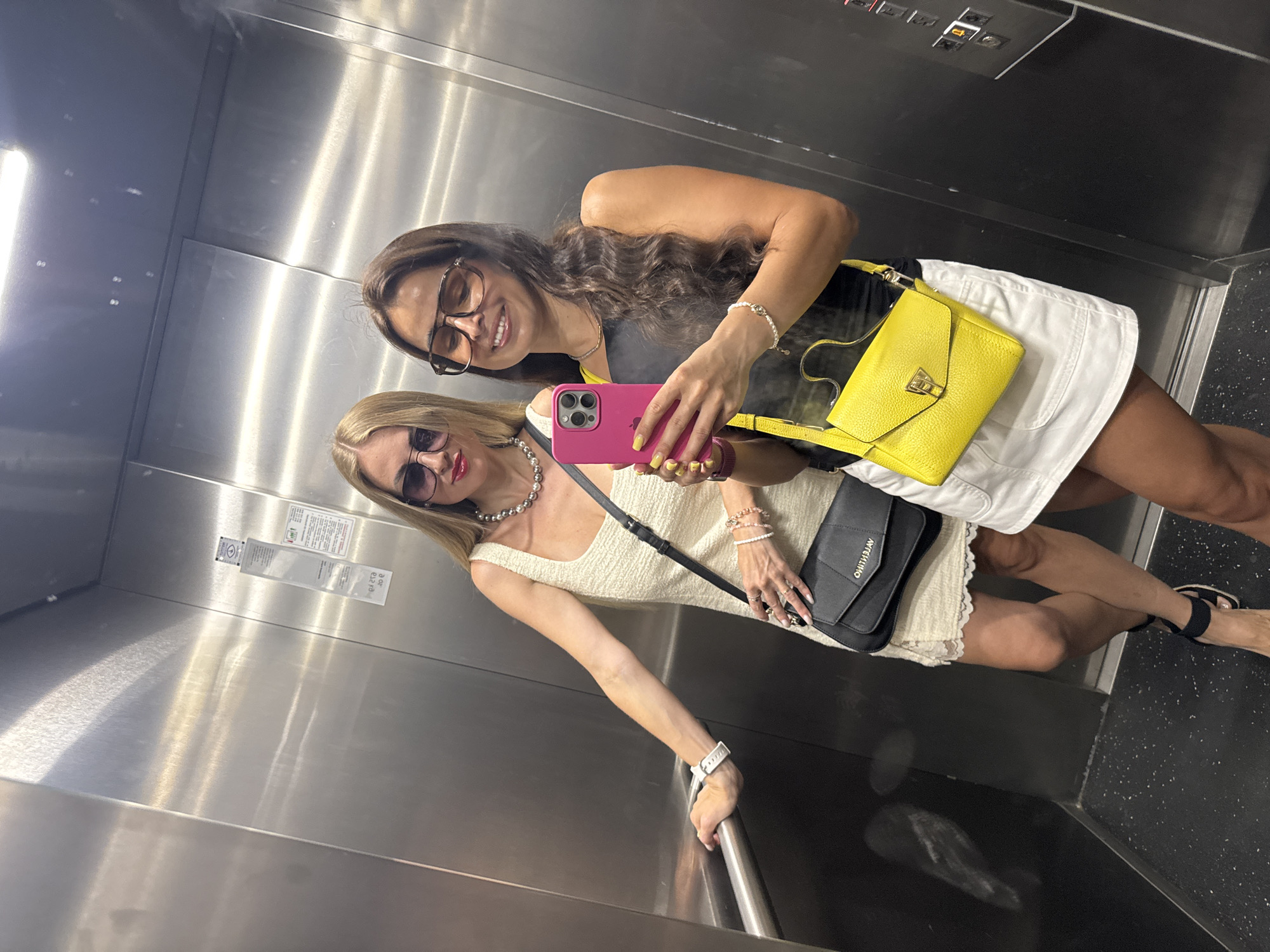 Two women are taking a selfie in an elevator. The woman on the left has blonde hair and is wearing sunglasses, a cream-colored dress, and a black crossbody bag. The woman on the right has long brown hair and is wearing sunglasses, a black top, a white skirt, and a yellow crossbody bag. She is holding a pink iPhone in her right hand and is smiling. The elevator walls are made of brushed metal, and there is a handrail on the left side.