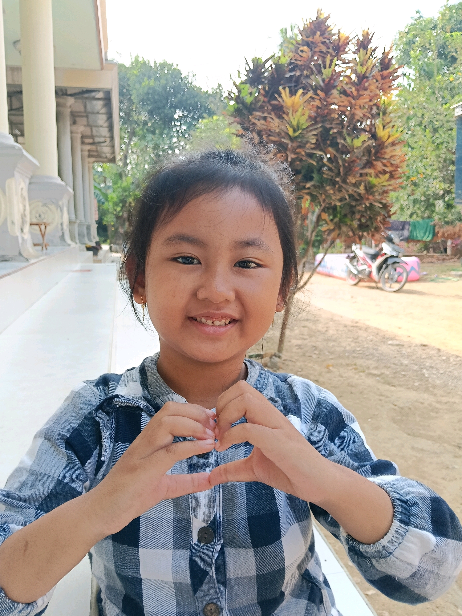 In an outdoor environment, a young girl is positioned front and center, her gaze directed forward and a smile gracing her face. She is forming a heart shape with her hands, which are brought together in front of her chest. Her attire consists of a blue and white checkered long-sleeved shirt. Her hair is dark and pulled back. The background of the image reveals a paved walkway, columns, and a building structure to the left. To the right, a plant with red and yellow leaves is visible, along with a motorcycle and a pink inflatable object. The ground appears to be dusty or sandy. The lighting suggests it is daytime.