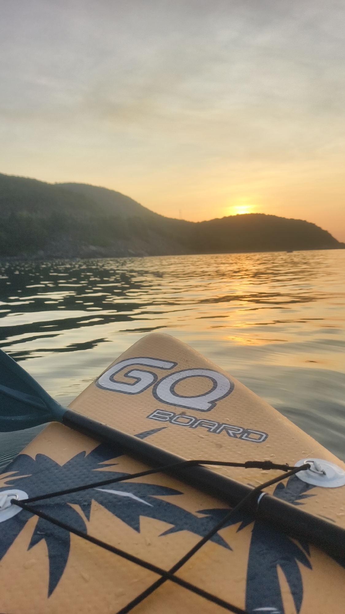 An eye-level, close-up shot captures the nose of a yellow paddleboard adorned with a palm tree design as it floats on calm, reflective water. The paddleboard has the text "GO BOARD" printed on it. A paddle blade, partially visible on the left, is in the water. In the background, a hazy silhouette of a lush, green island meets the horizon. The sky above is a muted blend of pale gray and soft yellow, with the sun setting behind the island, casting a warm glow and shimmering reflections on the water's surface. The scene is tranquil and serene, suggesting a moment of quiet enjoyment of nature.