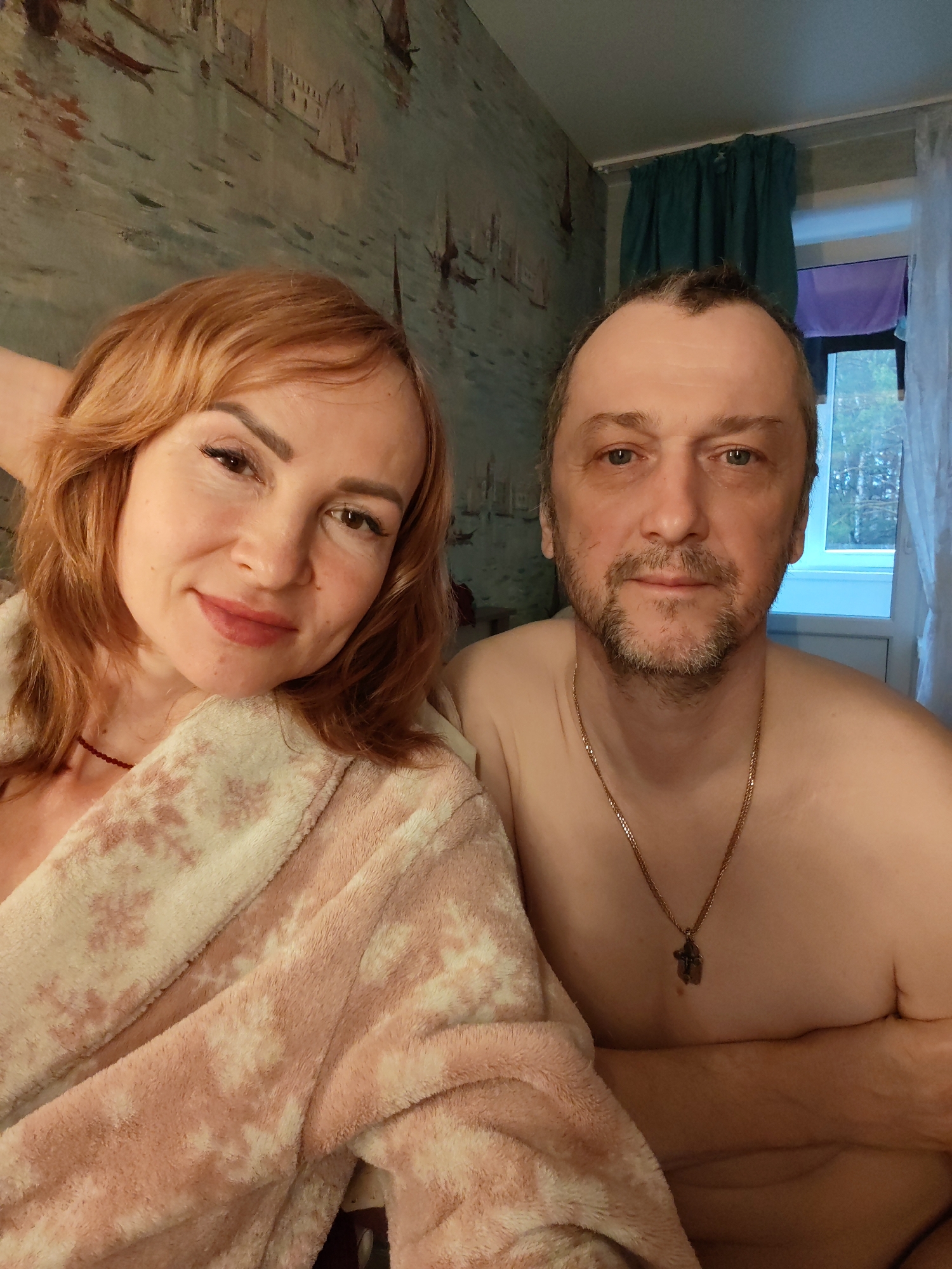 A close-up, eye-level shot depicts a middle-aged couple posing indoors. The woman, on the left, has red hair and is wearing a pink bathrobe. She is smiling and looking at the camera. The man, on the right, is shirtless, wearing a gold chain with a pendant, and also looking at the camera with a slight smile. The background includes a wall with a mural depicting a seascape, and a window with blue curtains and a purple sheet behind them. The setting appears to be a room in a private residence. There is no specific information linking the image to the city of Chaikovsky, Russia.