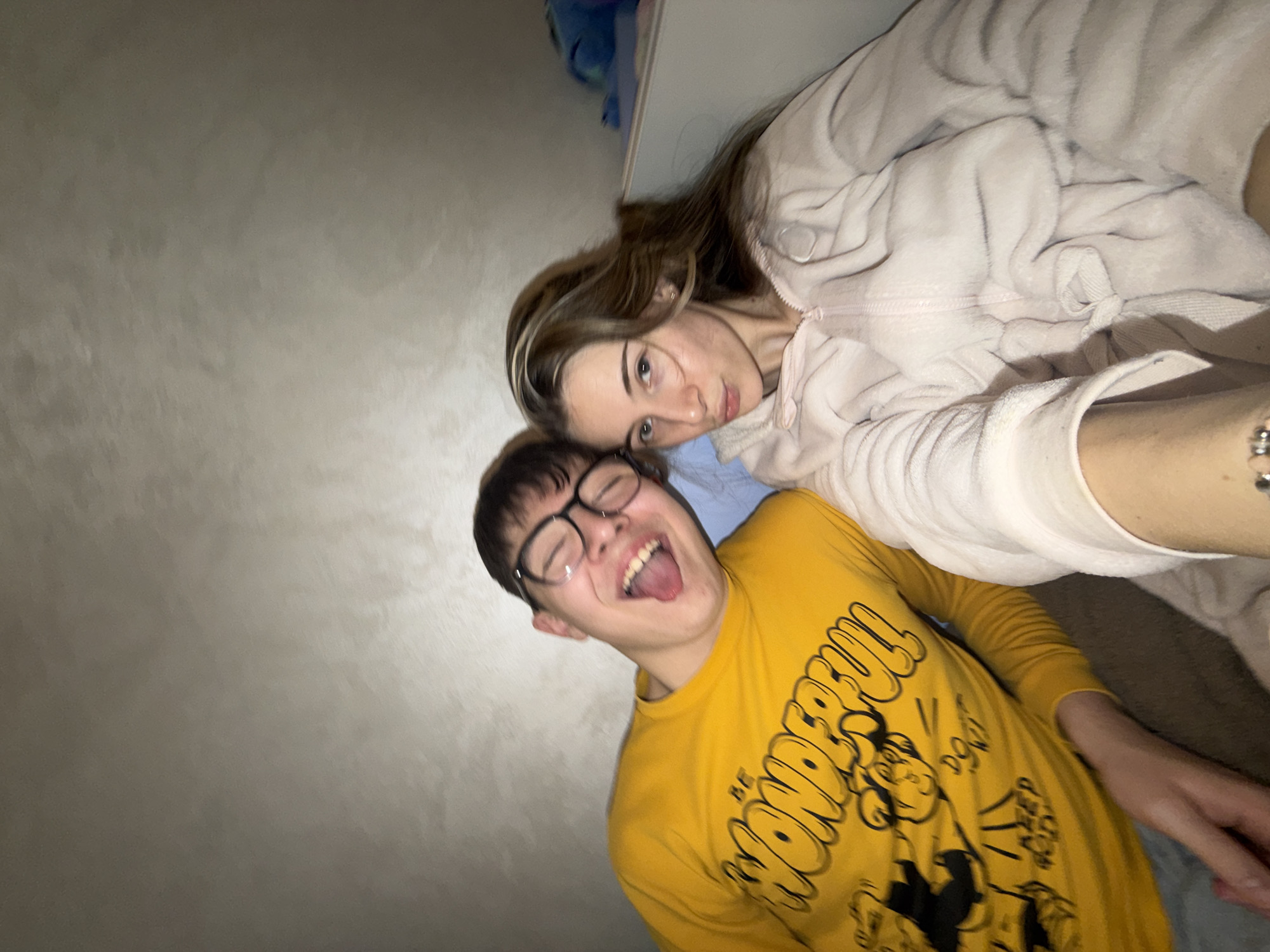 A photograph shows a woman and a man posing for a picture, likely in an indoor setting. The woman is positioned above the man, wearing a soft, light-colored hooded top. Her hair is long and brown, partially covering her shoulder. She is looking toward the camera. The man is wearing a yellow long-sleeved shirt with a black cartoon graphic. He is also wearing glasses and is sticking his tongue out. A gray wall is visible behind them. The photograph has a slightly tilted perspective. There is not enough information to locate the scene in Plovdiv, Bulgaria.