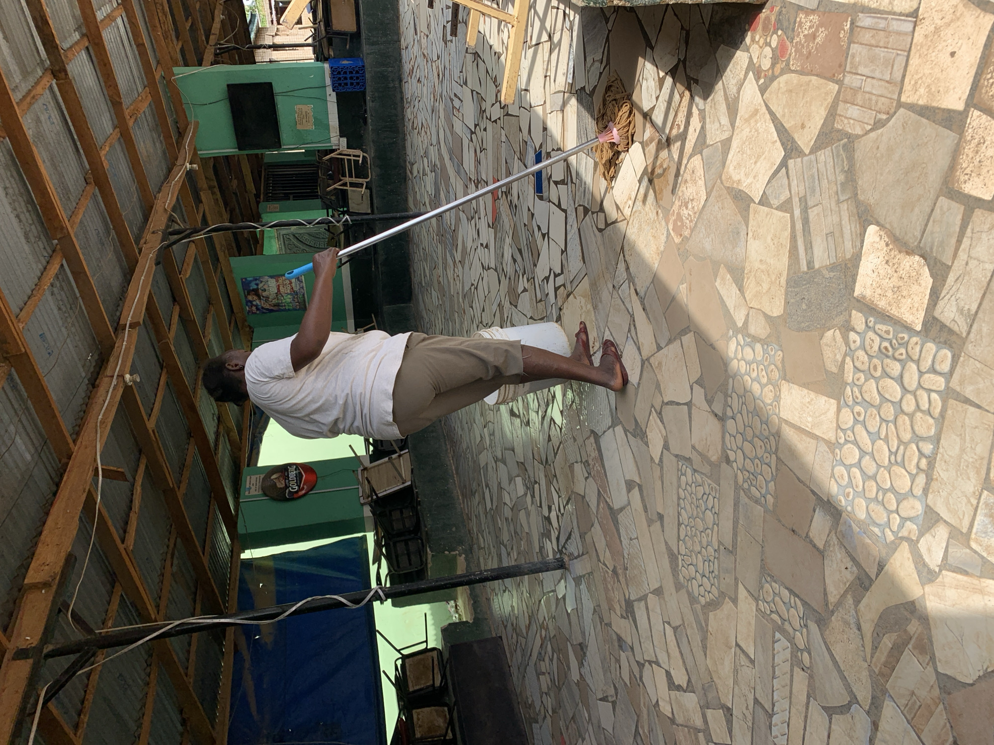 A man is mopping a mosaic-tiled floor in an outdoor setting near New Stadium, Nigeria. The man is wearing a white shirt and khaki shorts, and is barefoot. He is using a string mop and a white bucket. The environment includes a corrugated metal roof, a green building with a Goldberg beer sign, and several chairs stacked against the wall.