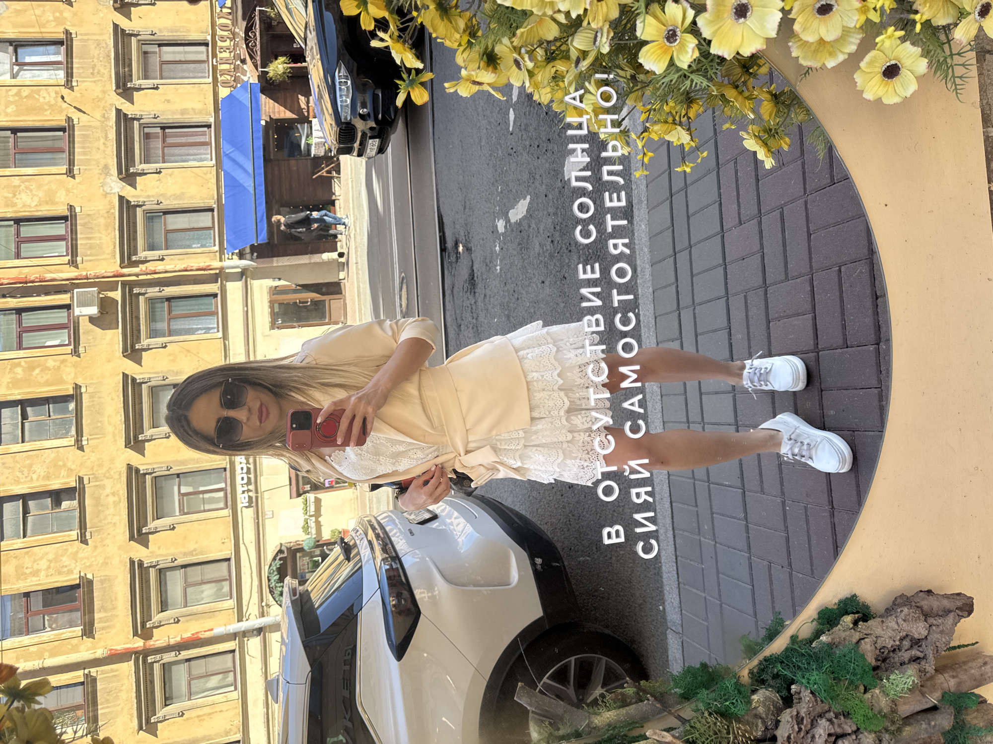 This is a vertical, slightly low-angle photograph taken in Saint Petersburg, Russia, featuring a woman taking a selfie in a large, oval mirror. The scene is outdoors, with a backdrop of a traditional yellow, multi-story building with many windows. The weather appears to be sunny, indicated by the bright lighting and shadows.
The woman is the main subject, and she is wearing a pale yellow blazer over a white lace dress. She has on sunglasses and is holding a red smartphone to take the picture. Her legs are visible, she is wearing white sneakers, and she is standing next to a white SUV reflected in the mirror.
The mirror itself is a prominent object, framed by a decorative arrangement of yellow flowers and greenery. The reflection in the mirror shows the woman, her attire, the white SUV, and an urban setting with a paved sidewalk and a street. The reflection also reveals text written in white, bold, Cyrillic letters on the mirror's surface, which reads "В ОТСУТСТВИЕ СОЛНЦА! СИЯЙ САМОСТОЯТЕЛЬНО!" which translates to "IN THE ABSENCE OF SUN! SHINE ON YOUR OWN!". The text is positioned vertically within the mirror's reflection.
The scene suggests a casual outing on a pleasant day. The architecture of the building and the presence of a car on the street are typical of an urban environment. The woman's pose and the selfie suggest she is documenting her appearance and perhaps enjoying her time.